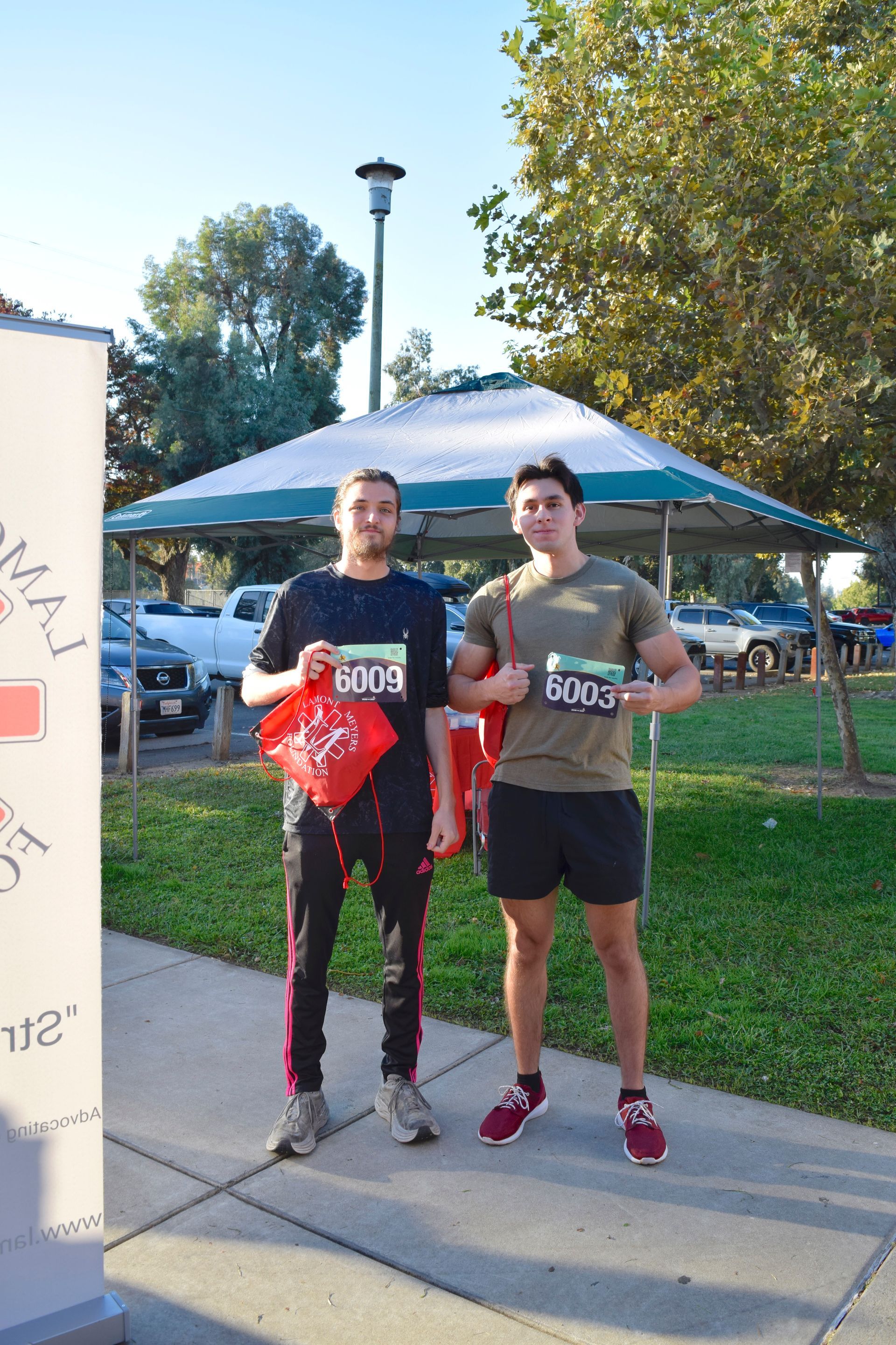 Two runners pose outdoors after race; one in black sweats, the other in athletic shorts, holding race bibs.