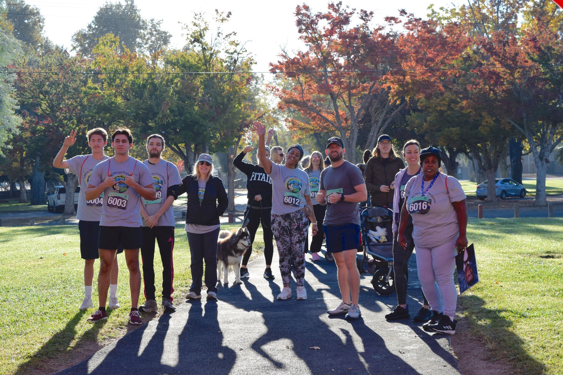 Group of people on a path, some wearing race bibs, waving. Trees in the background. Sunny day.