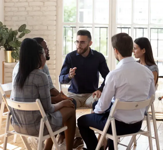 Group therapy session in progress; people seated in a circle, one person speaking and gesturing. Bright, natural light.