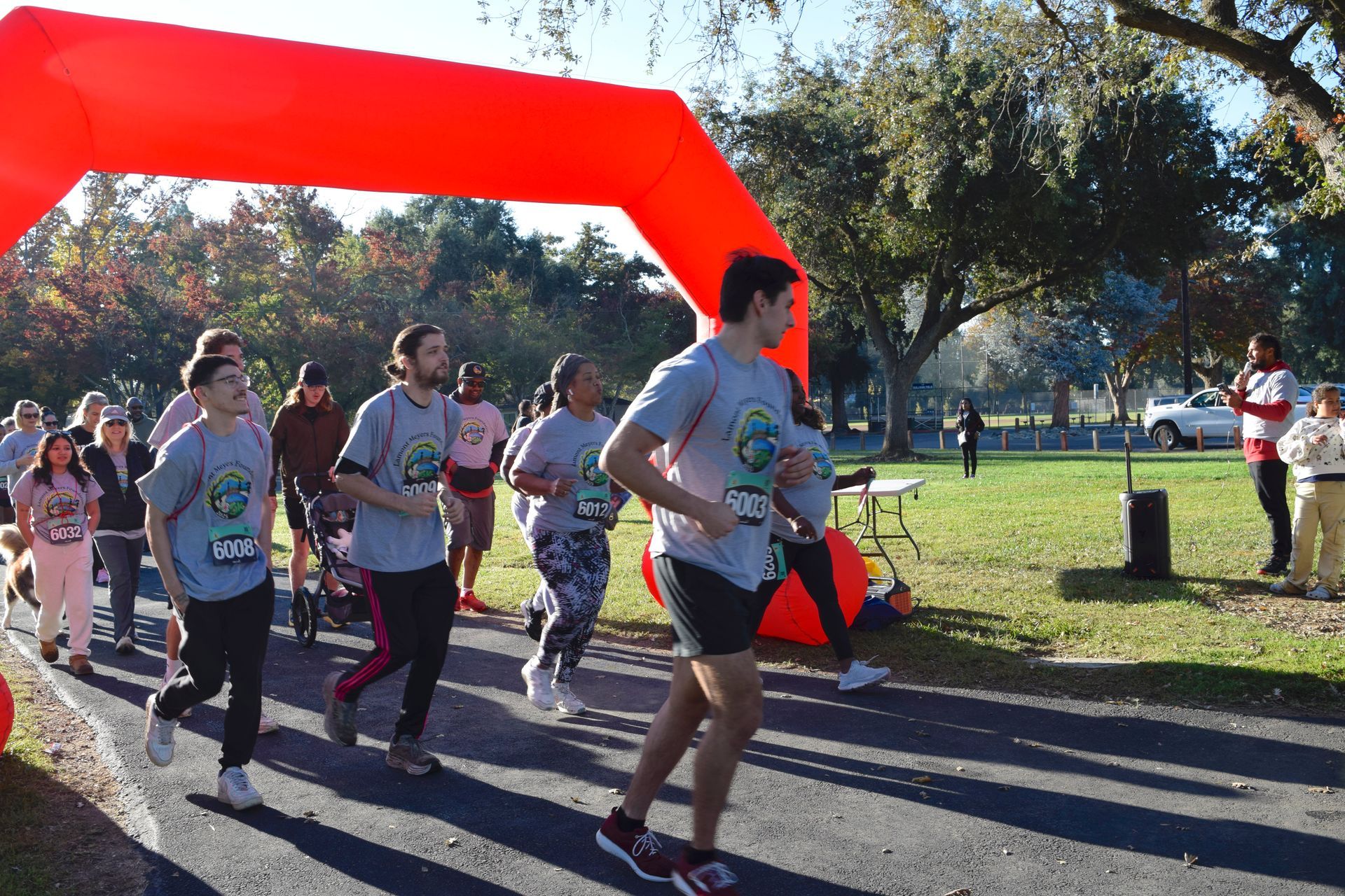 Runners in a race under a red arch, near a park with spectators and announcer.