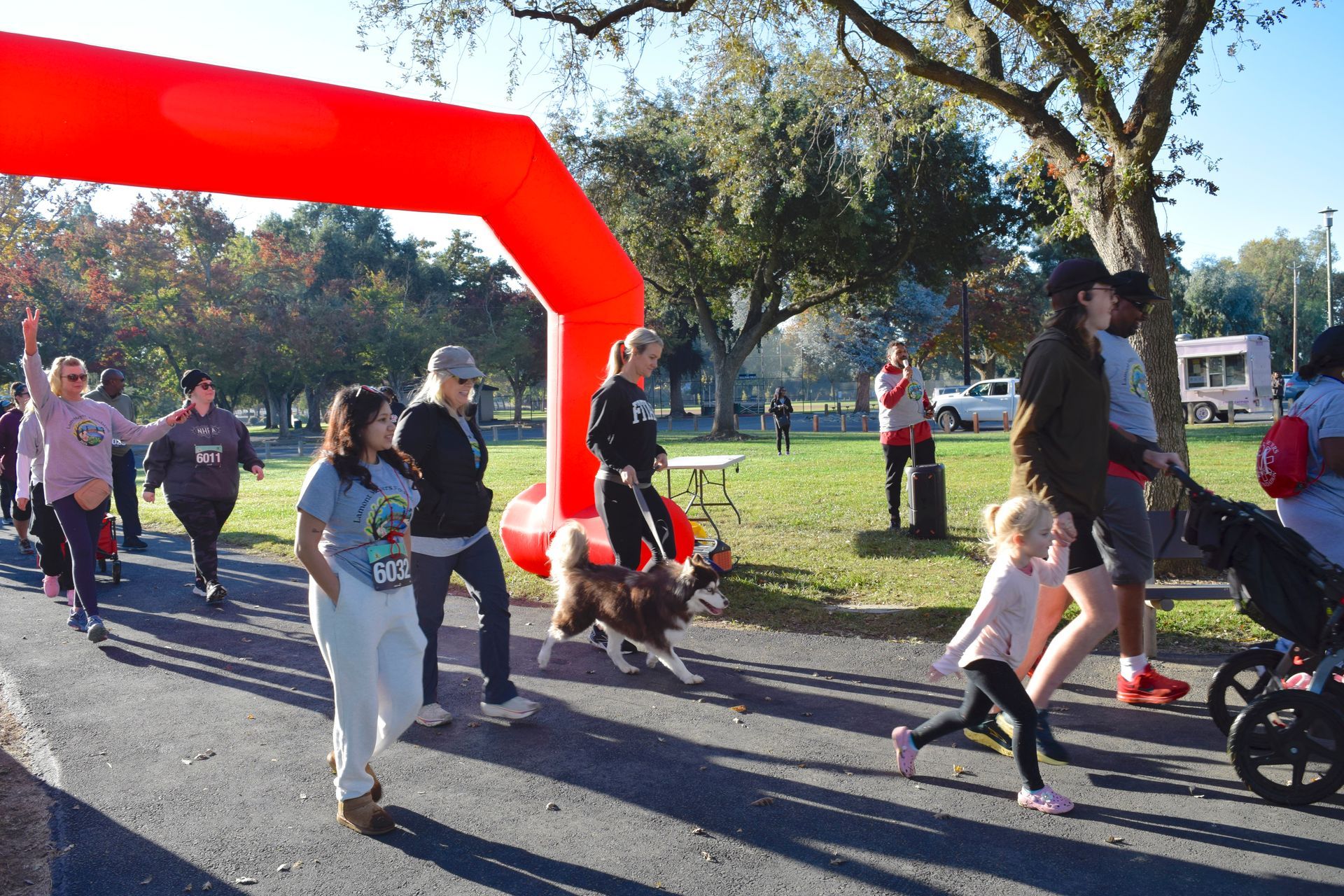 People walking and running under a red arch during an outdoor event in a park, including a dog and a stroller.