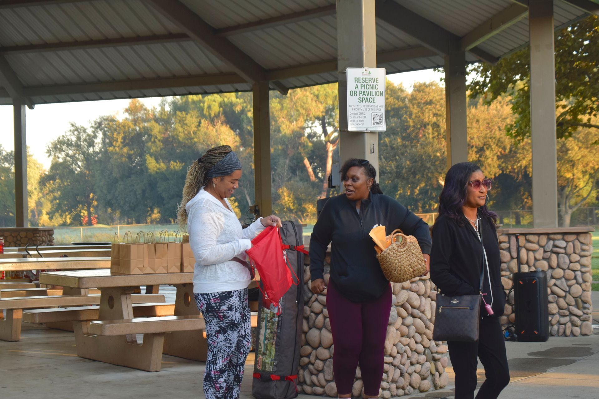 Three people at a picnic shelter. One opens a red bag, others hold baskets and purses. Trees in background.