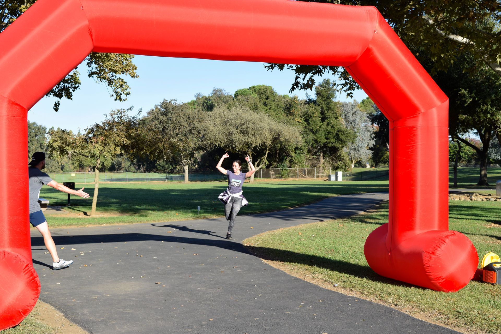 Runner crossing a red inflatable finish line arch on a paved path in a park; a person cheers.