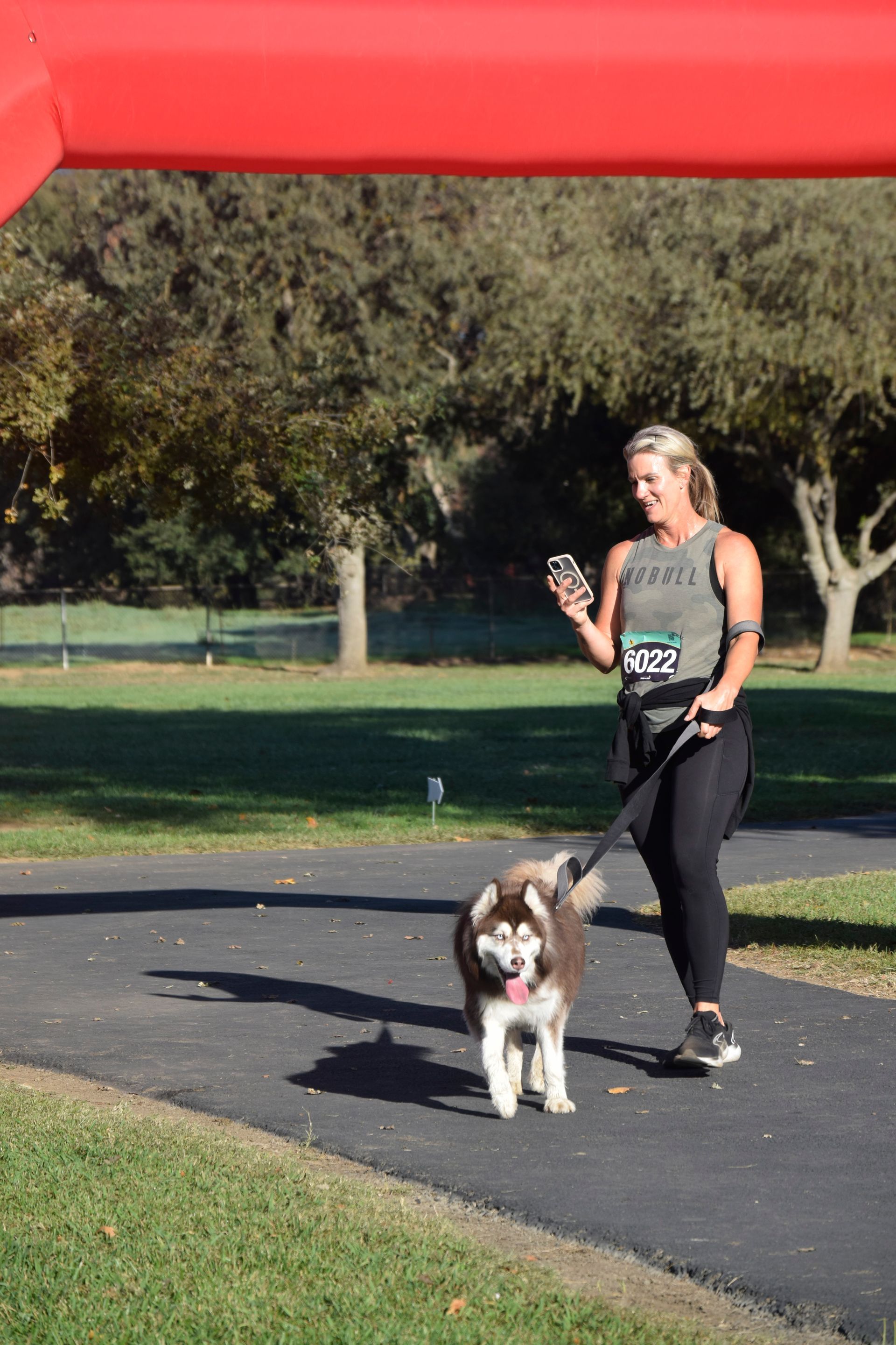 Woman and husky dog crossing finish line in park. Woman holds phone, dog on leash; red arch in background.