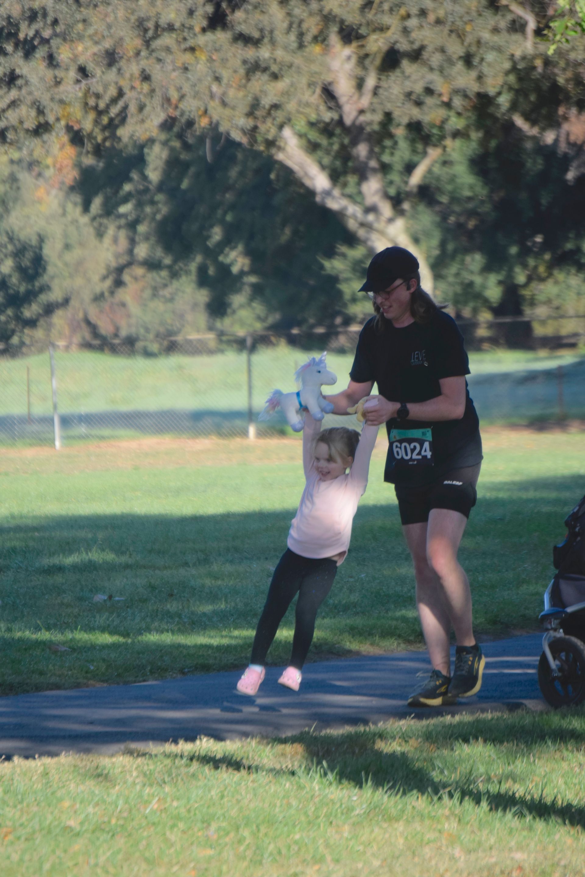 Woman in athletic wear swinging a child holding a stuffed animal on a park path.
