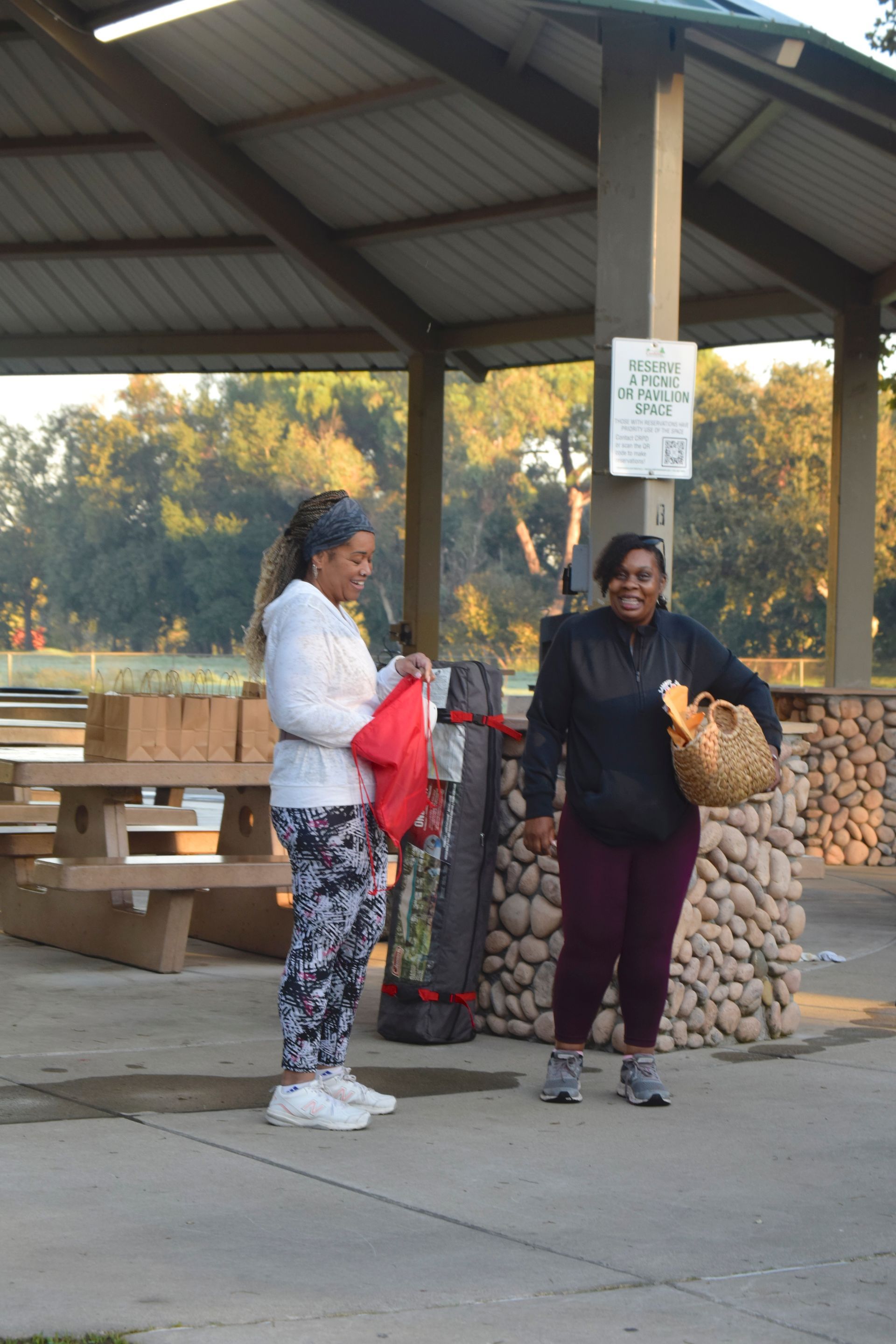 Two women at a park shelter smiling, one holding a red bag and the other a basket.
