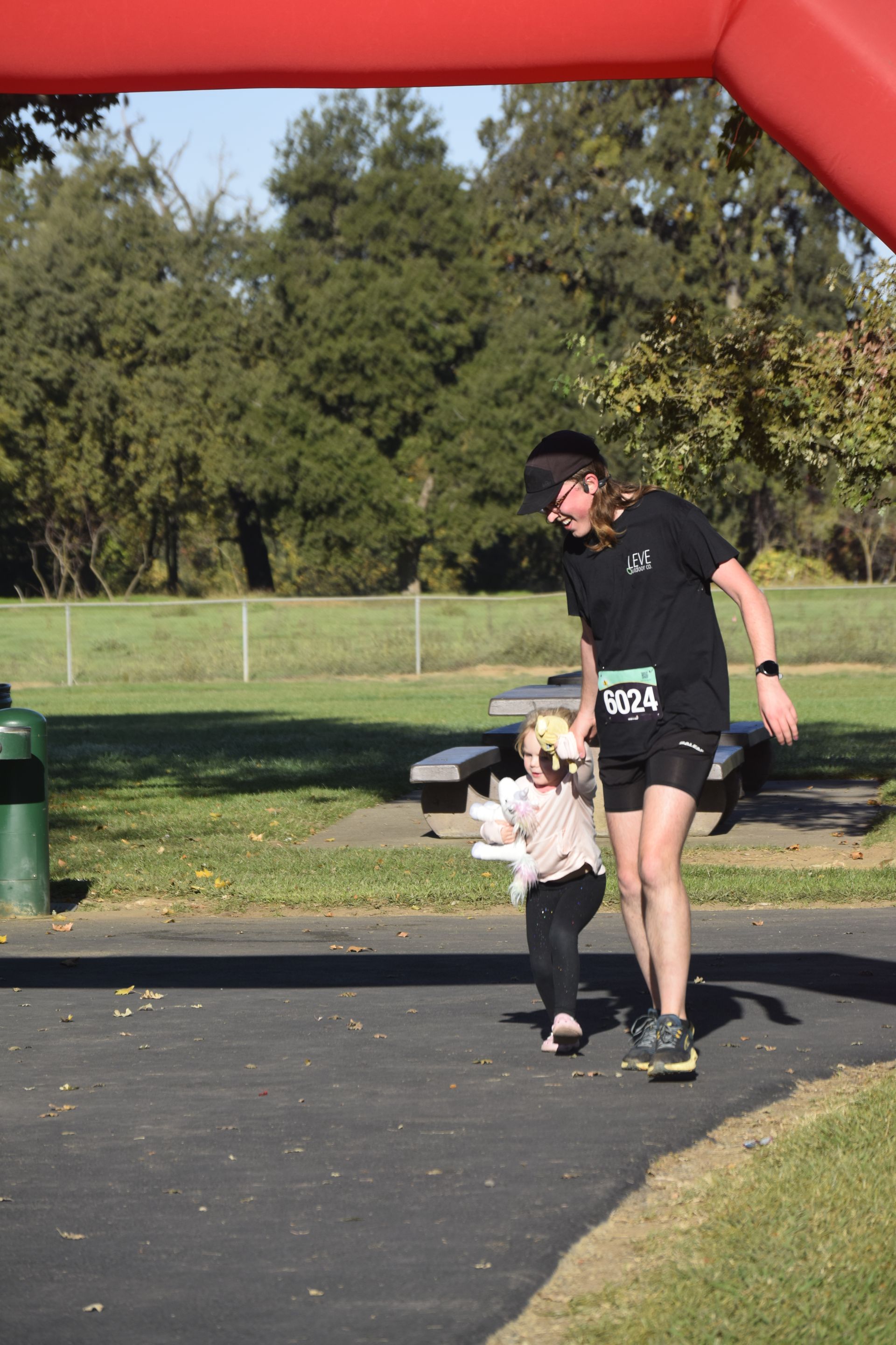 Man crossing finish line with a small child. They are running on a paved path in a park under a red arch.