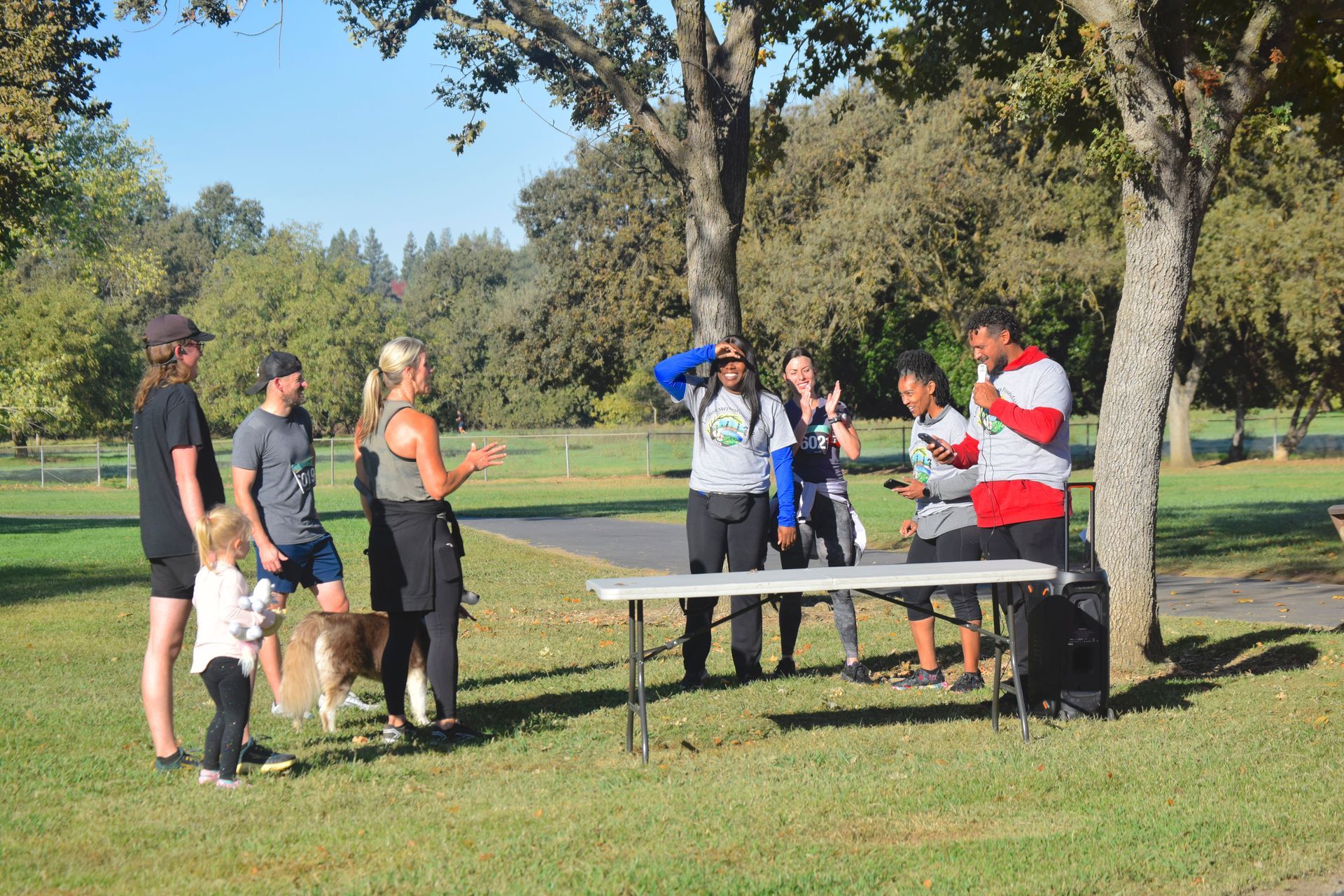 People at a park, near a table. Some are talking, others look on. Sunny day.