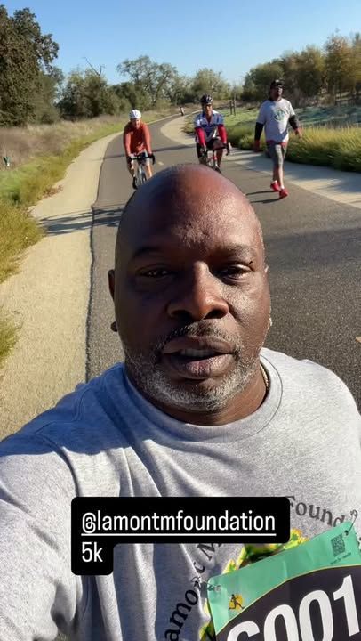 Man taking selfie on paved path, cyclists and walker in background. Sunny day, outdoor setting.