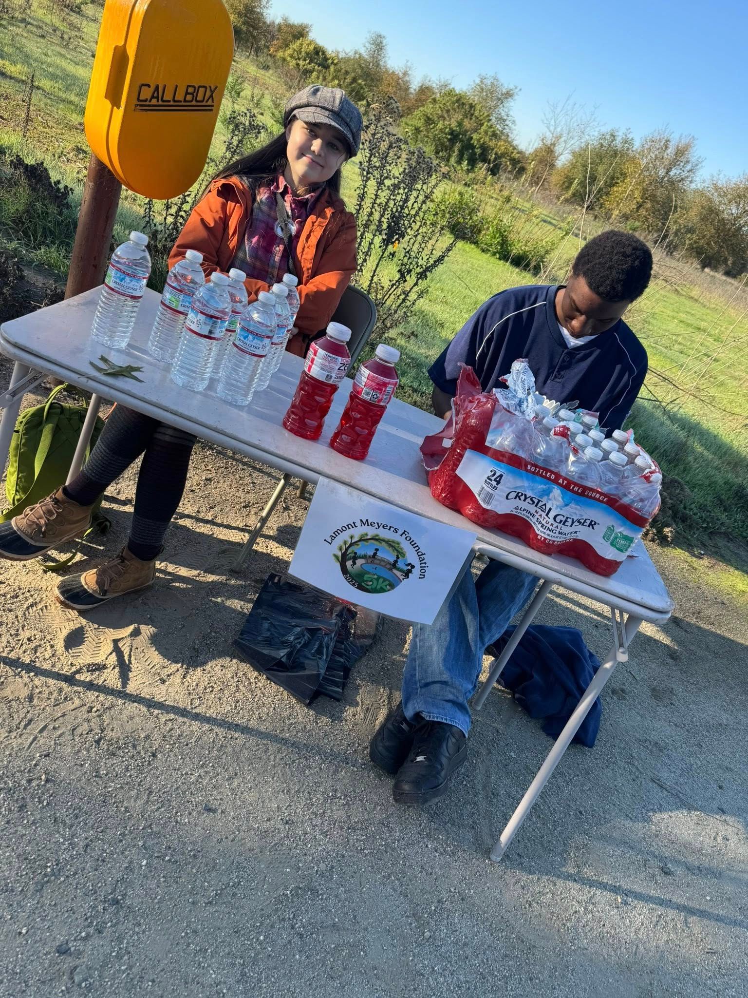 Two people at a table selling water bottles outdoors. A sign with a logo is visible.
