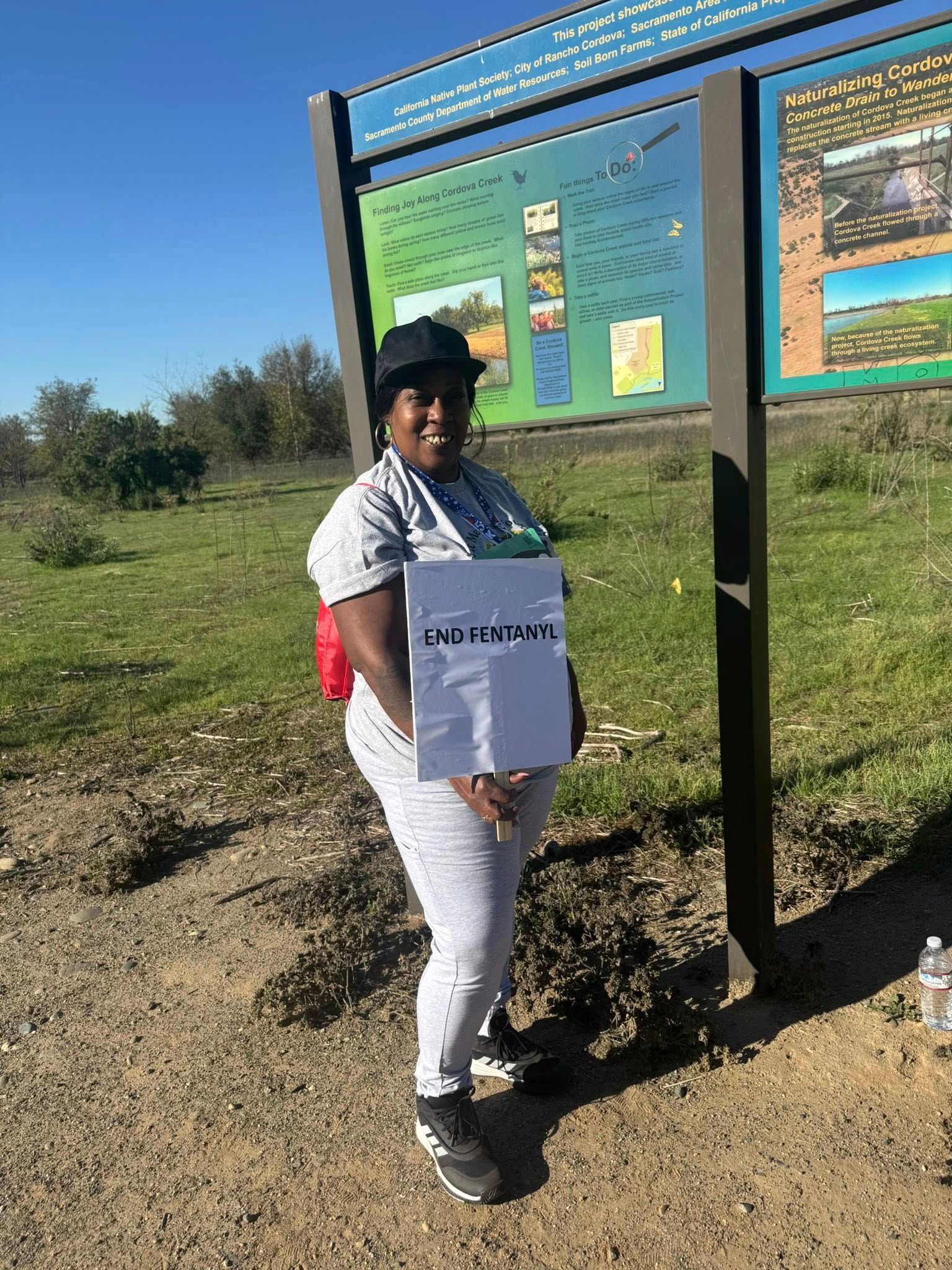Person standing by a park sign, holding a white sign. They are on a dirt path with green vegetation and a blue sky.