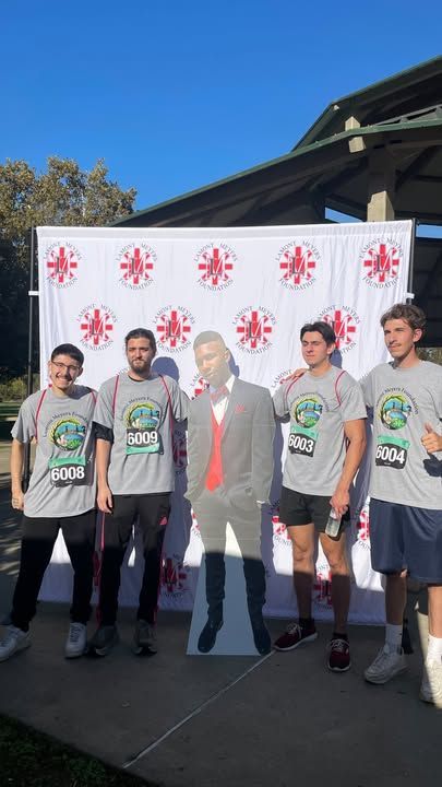 Five people pose with a cardboard cutout at a race backdrop. They wear race bibs and gray shirts.