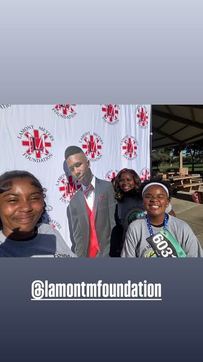Three people pose with a cutout. Two smile; one wears a bib. Background is a shelter with picnic tables.