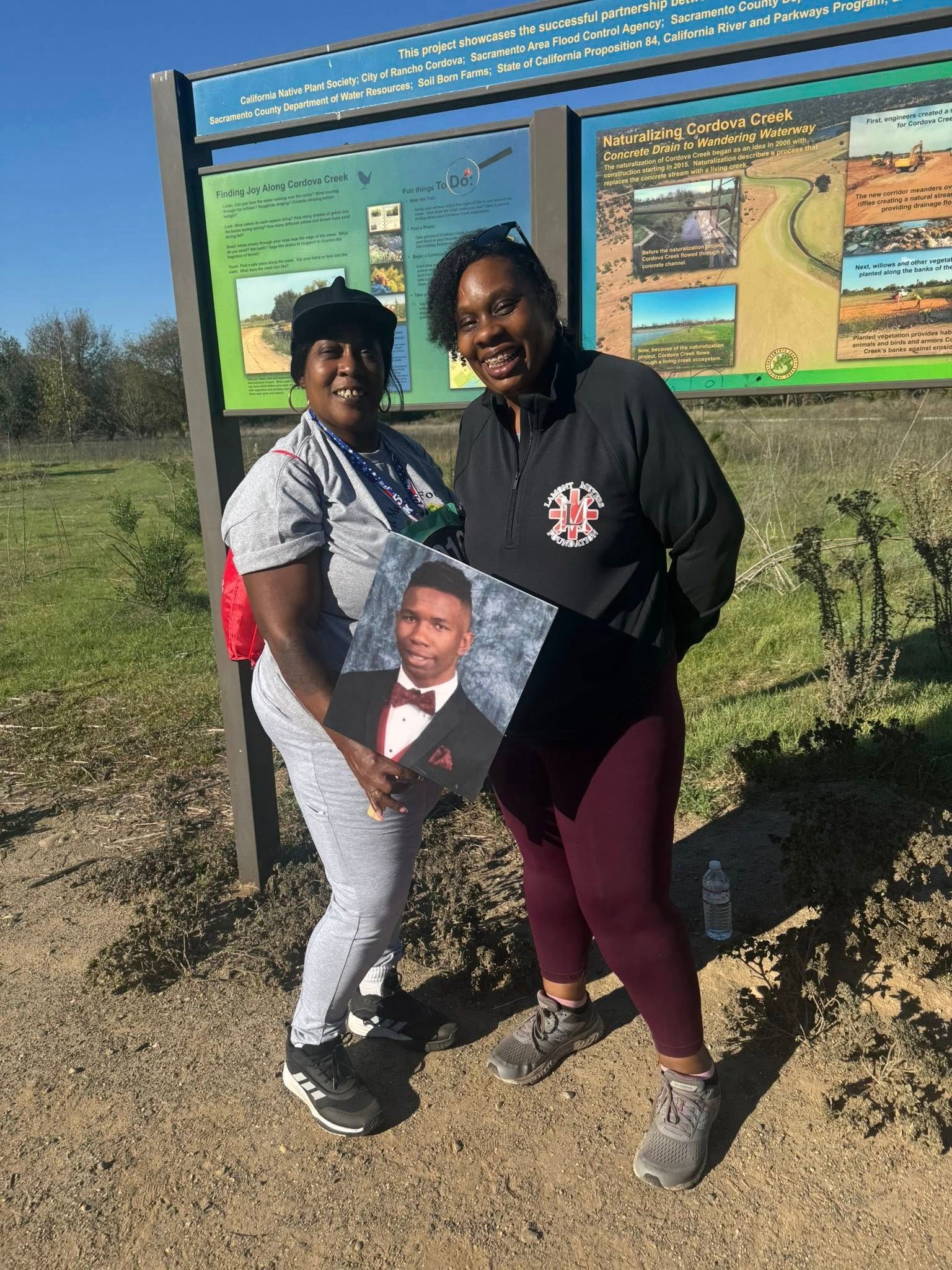 Two women holding a photo of a young person, smiling, near a park sign.
