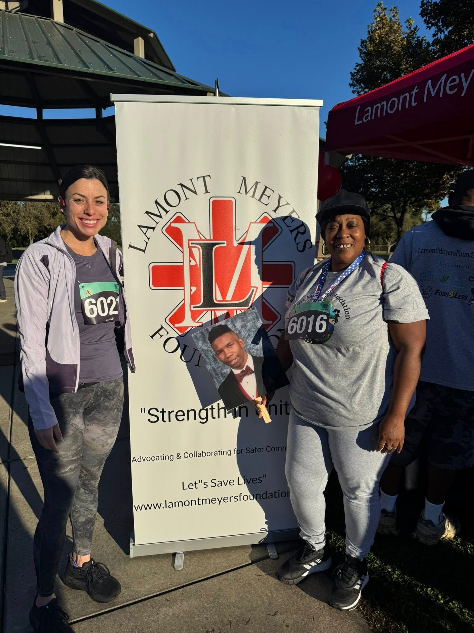 Two people pose next to a banner with a logo. One holds a photo of a man. Outdoor setting, sunny.