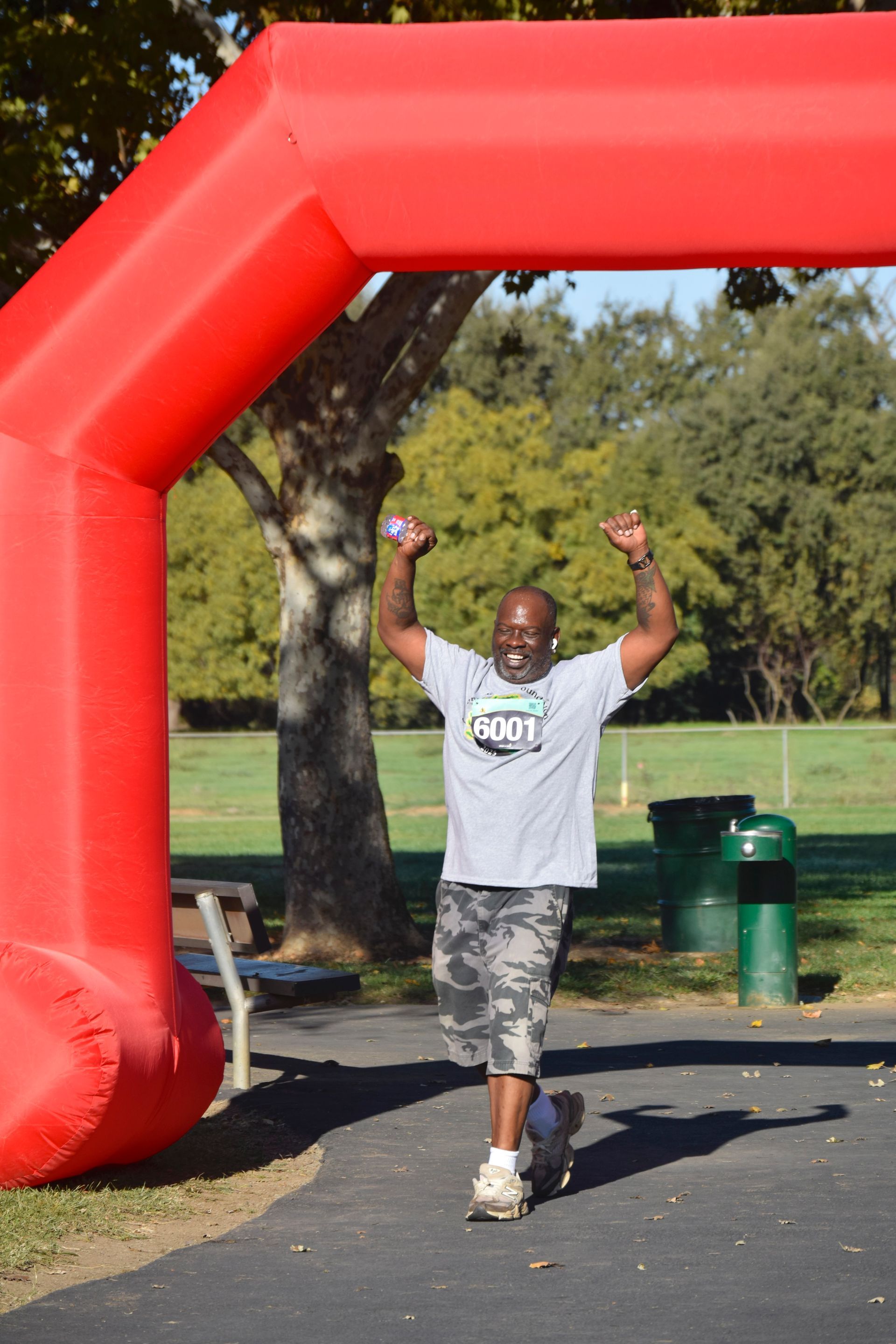 Man crosses finish line with arms raised under red arch. Park setting.