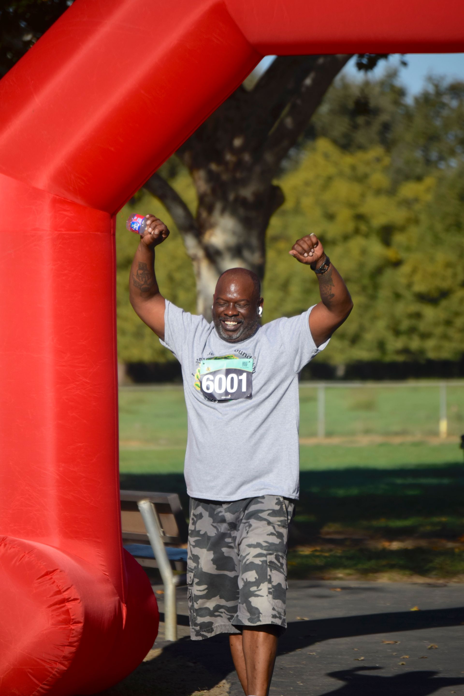 Man with arms raised, crossing a red finish line arch in a park, smiling.