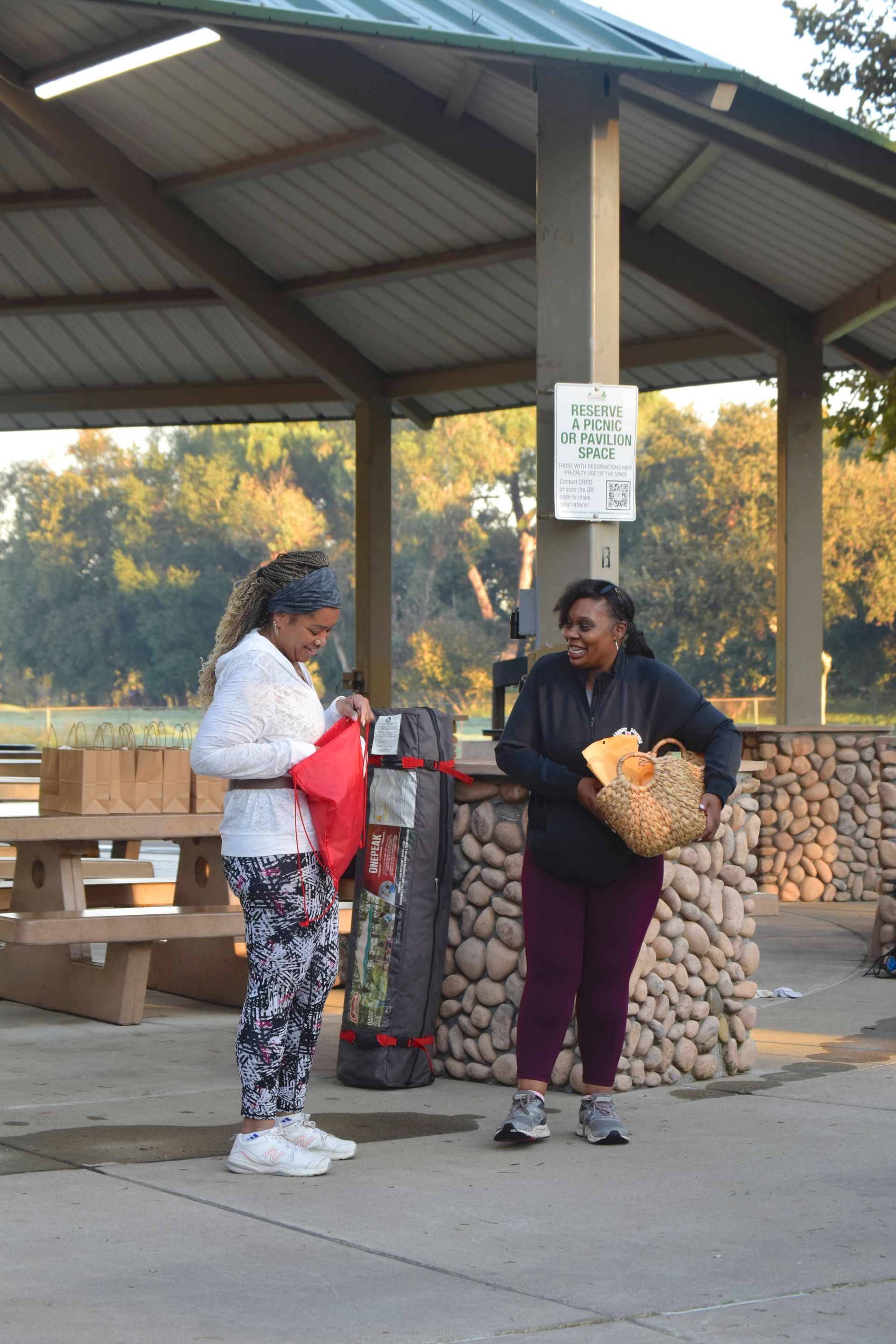Two people near a picnic shelter; one holds a basket, the other a red bag.