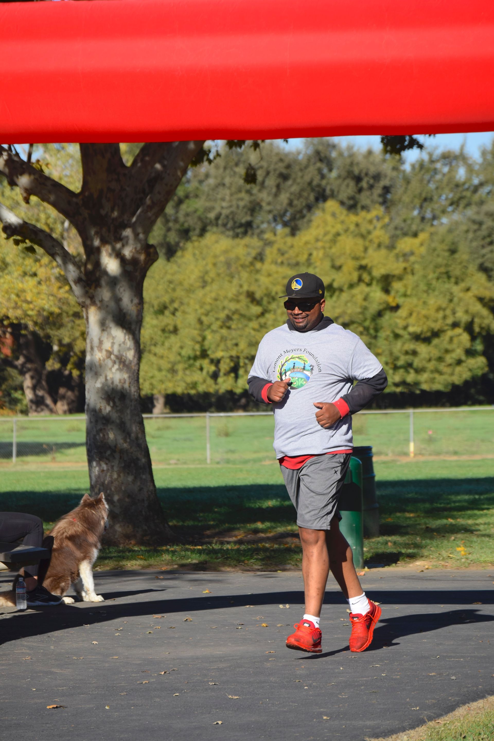 Runner in gray shirt and red shoes passing a finish line. Dog watches nearby.