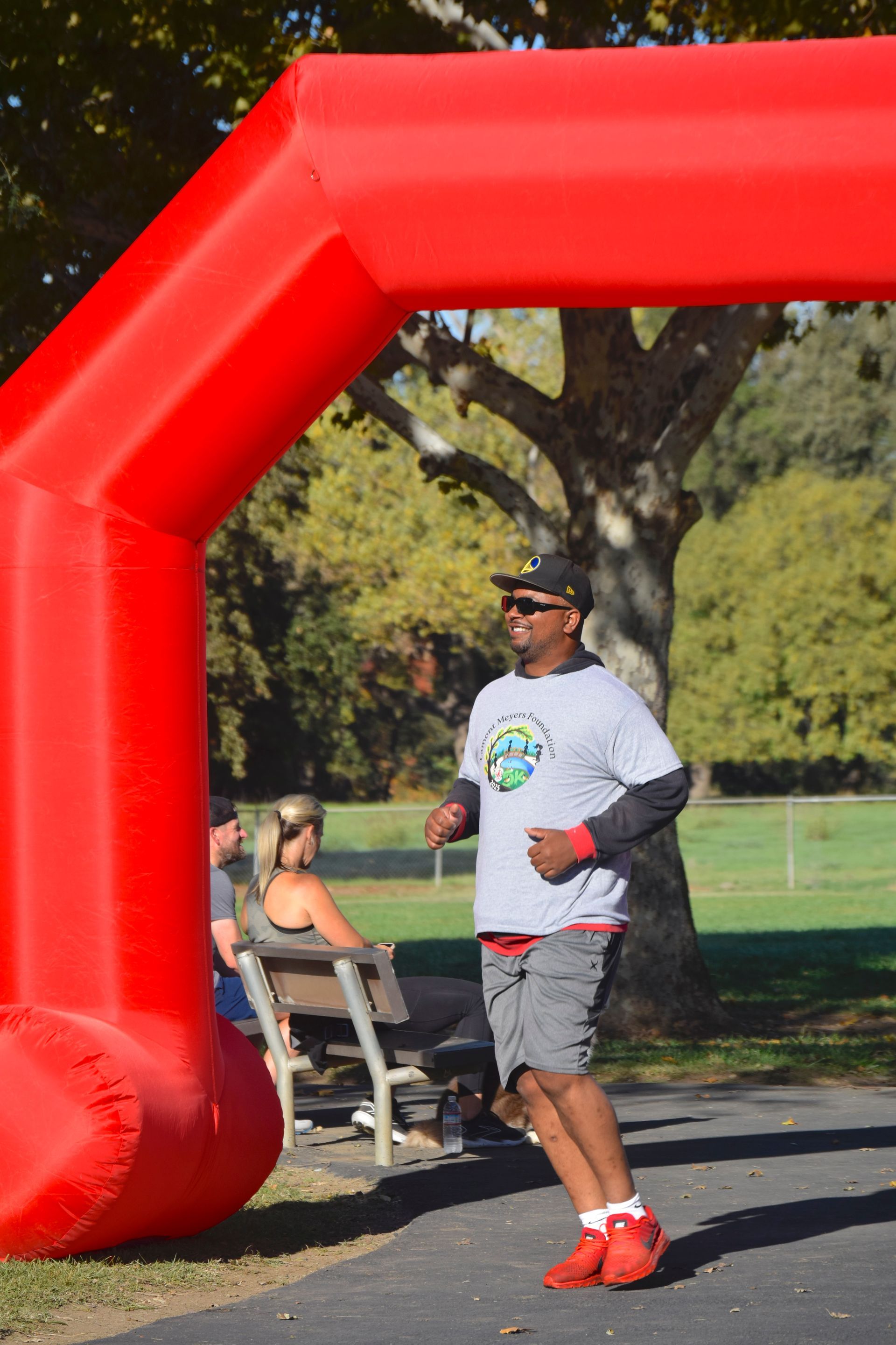 Man running toward the finish line arch. He is wearing a gray shirt, red shorts and shoes. Park setting.