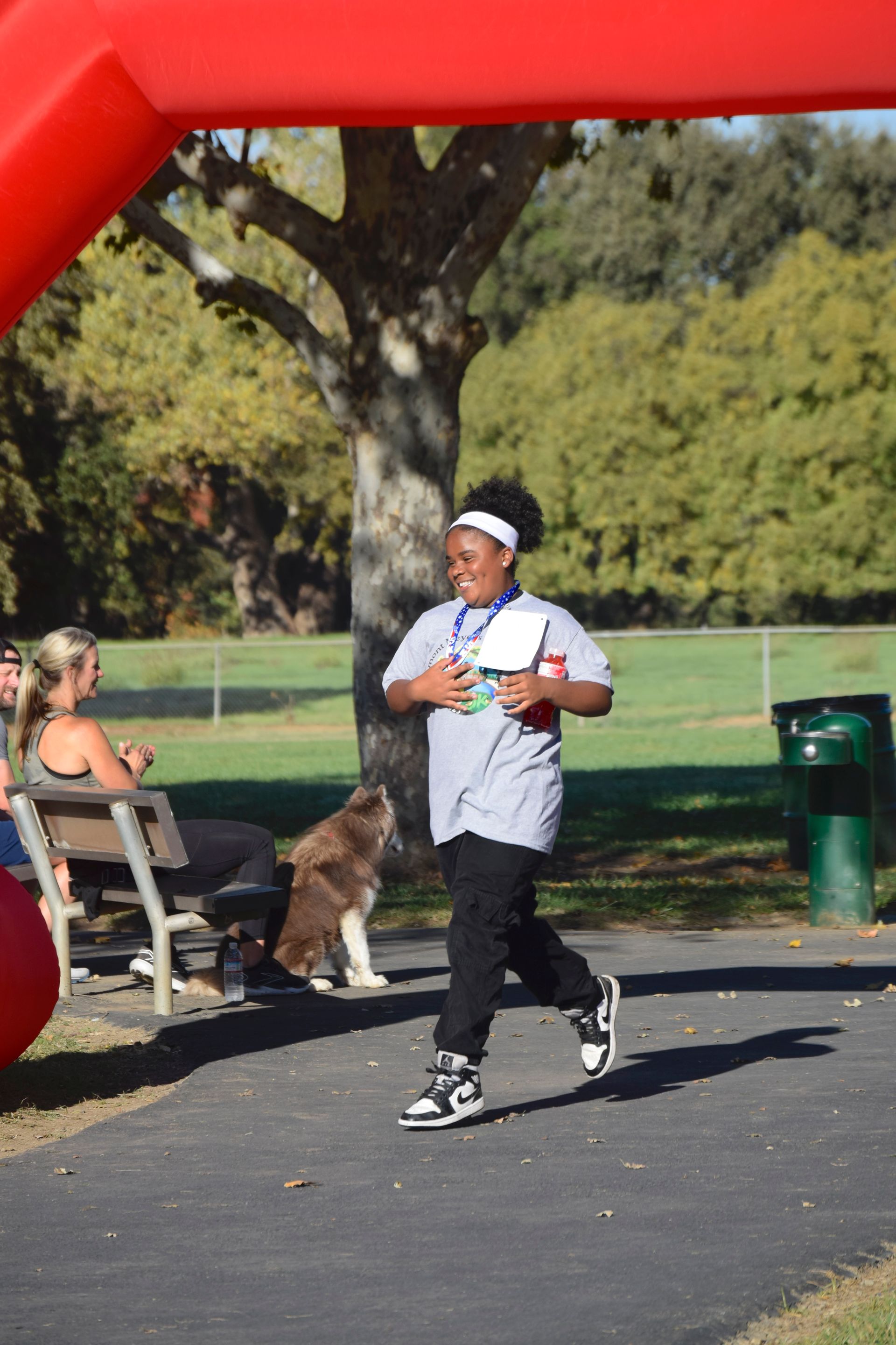 Person running towards the finish line under a red archway in a park, wearing a medal.
