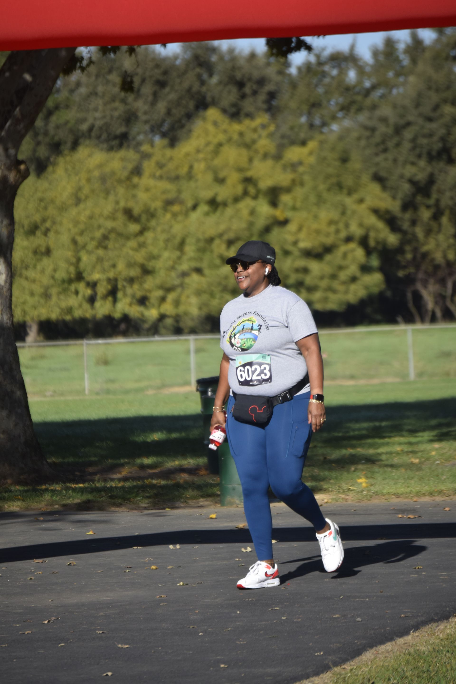 Woman in athletic wear walks on path under a red banner. Wearing a hat, glasses, and carrying a water bottle.