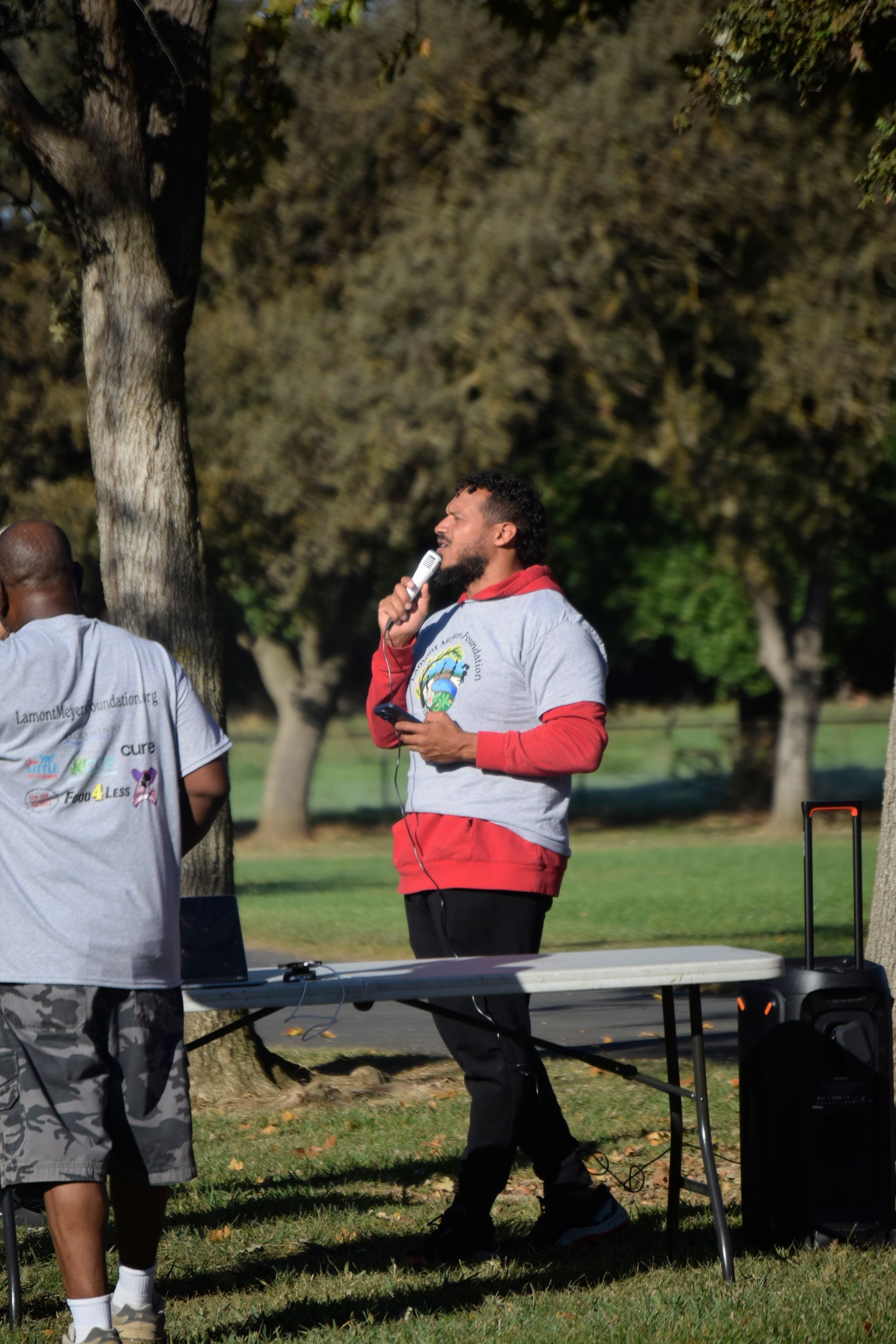 Man speaking into a microphone at an outdoor event, standing behind a table, with another person nearby.