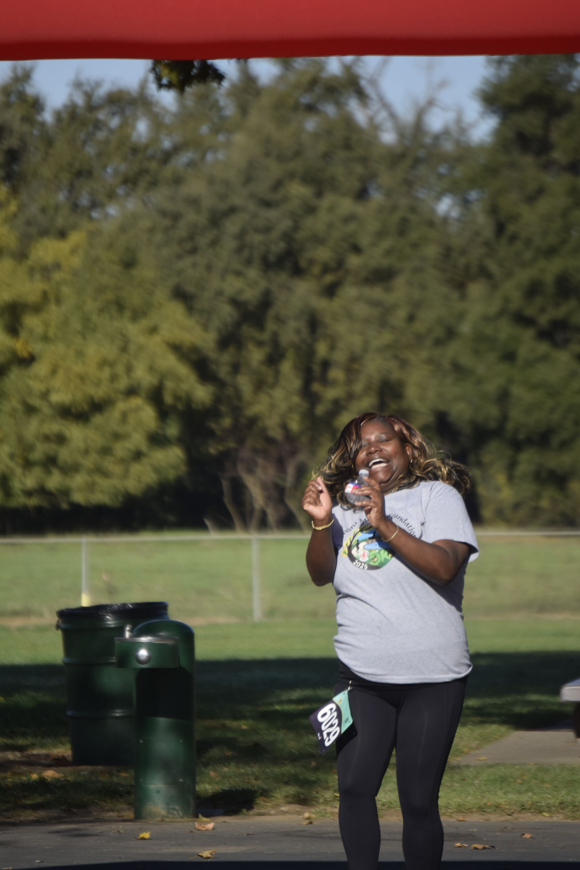 Woman in gray shirt and black leggings, smiling and gesturing, in park with green trees and trash cans.