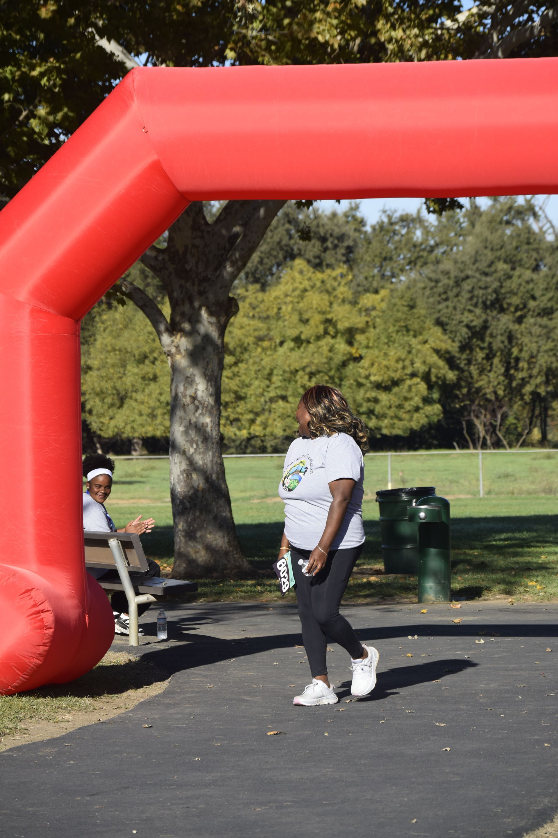 Woman walking under a red inflatable arch in a park, another person seated nearby.