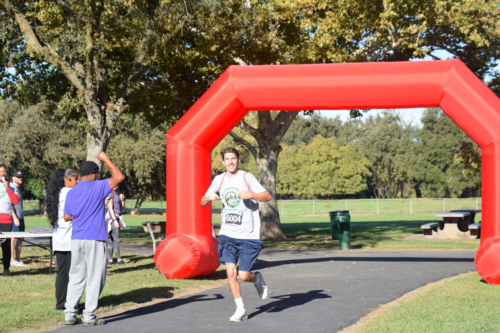 Runner crossing a red inflatable finish line in a park. Person with bib number, smiling, arms up.