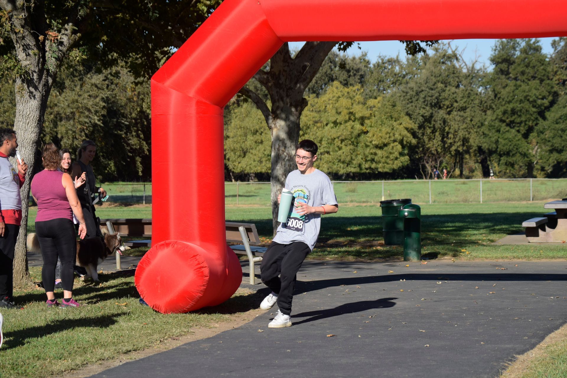 Young person crosses finish line under red arch, holding trophy, park setting.