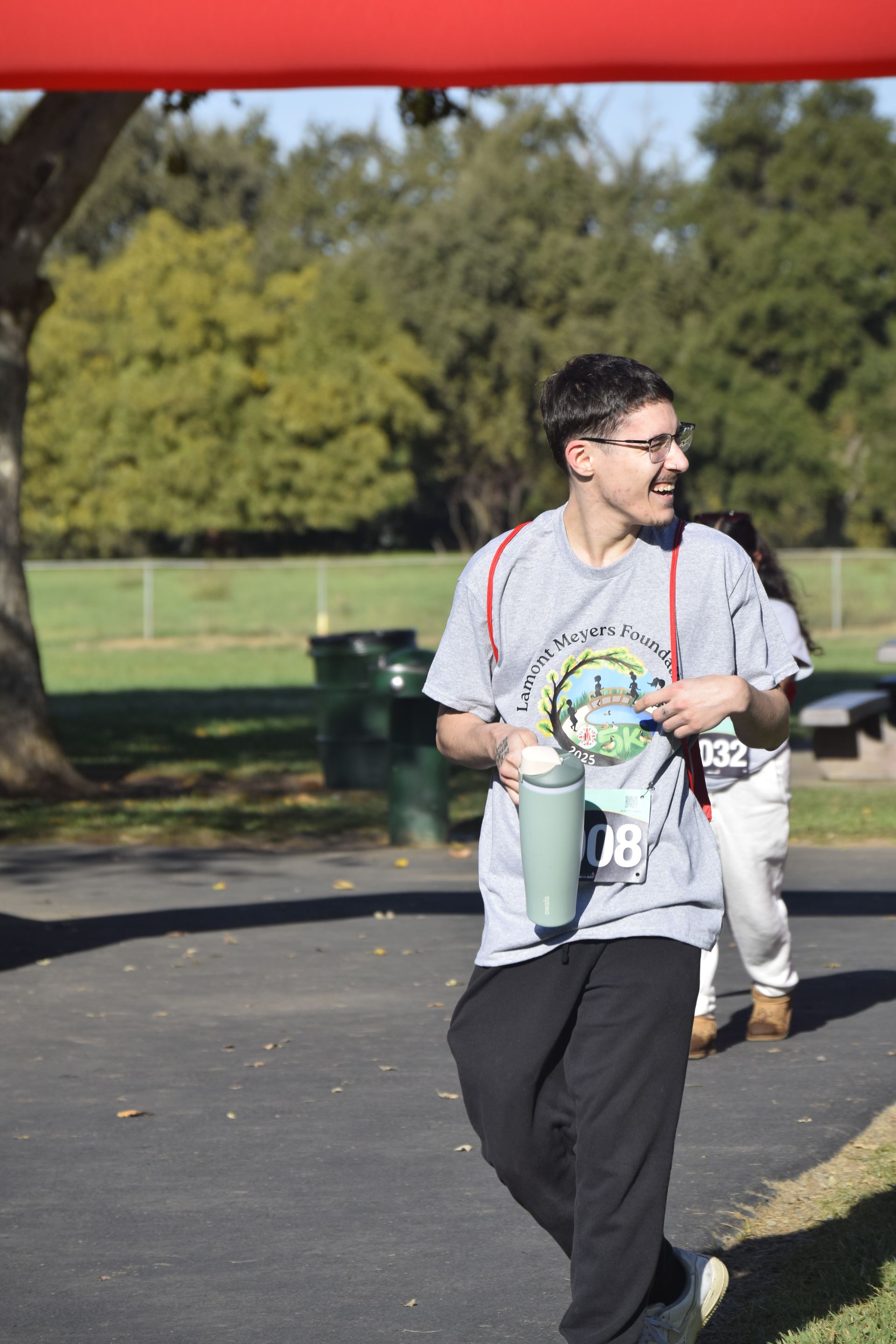 Person in gray shirt with race bib and cup, smiling, at an outdoor event near trees.