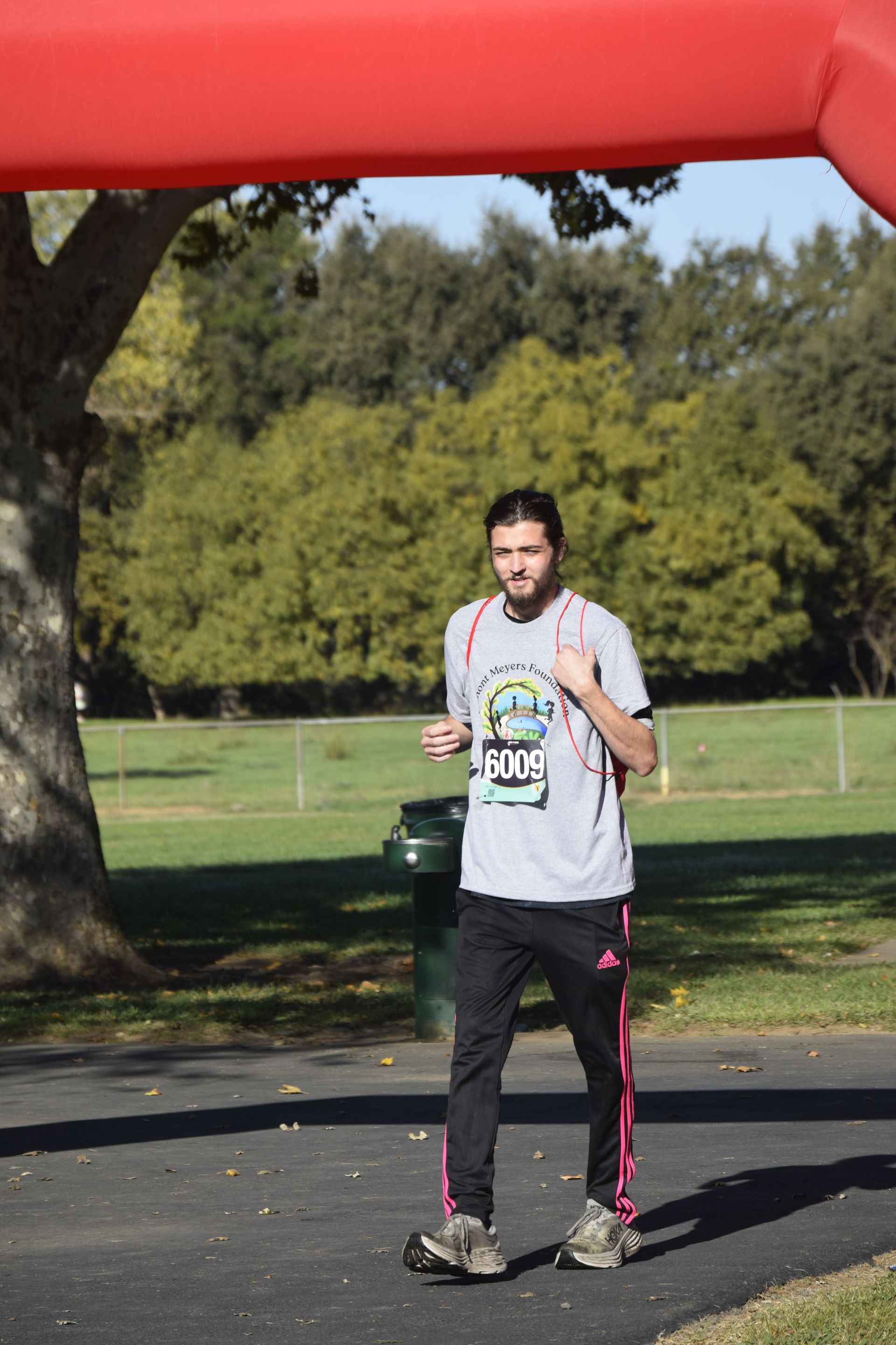 Man running under a red archway in a park, wearing a gray t-shirt, black pants with pink stripes, and running shoes.