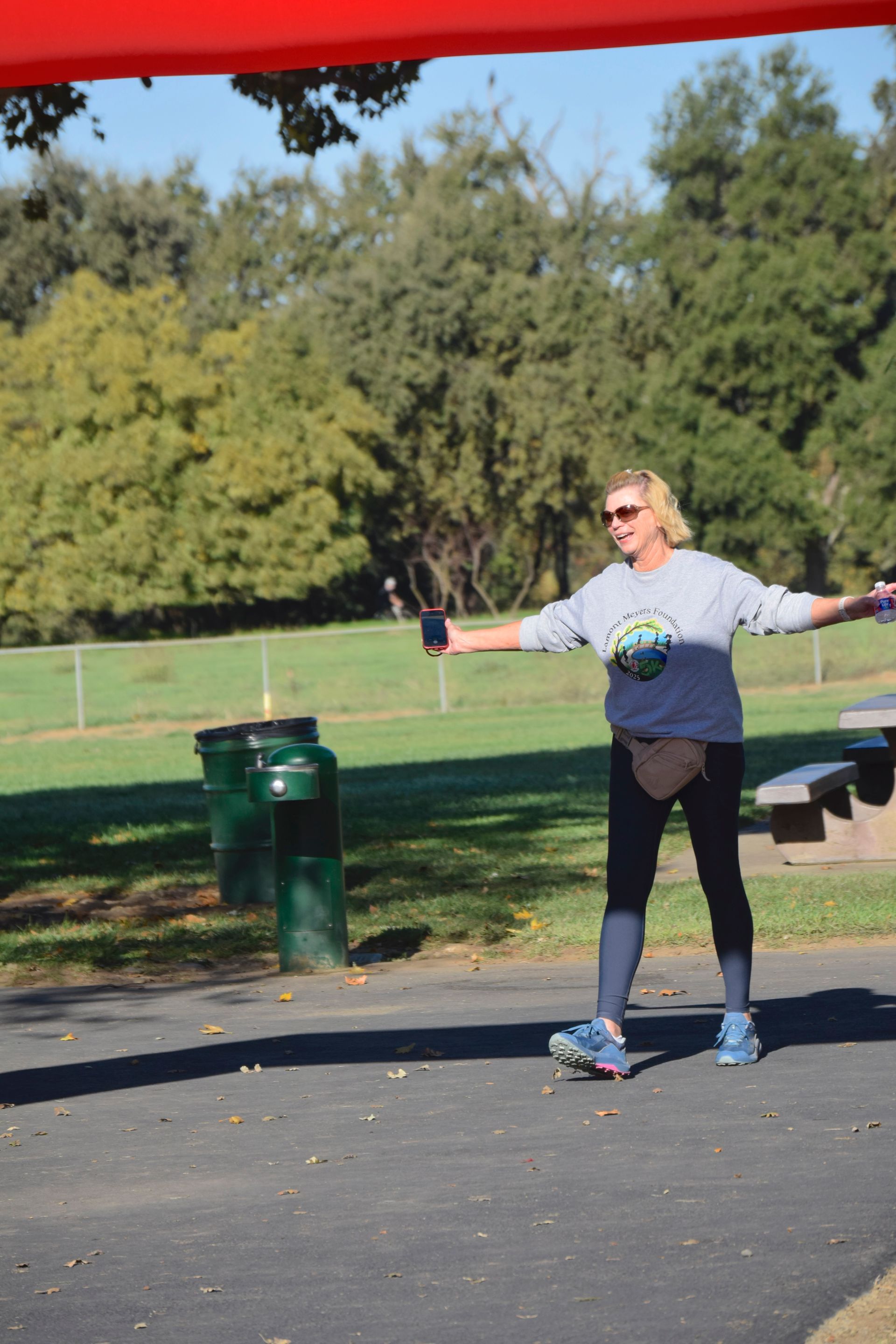 Woman outdoors with arms outstretched, holding phone. Park setting with trees, trash cans, and picnic tables.