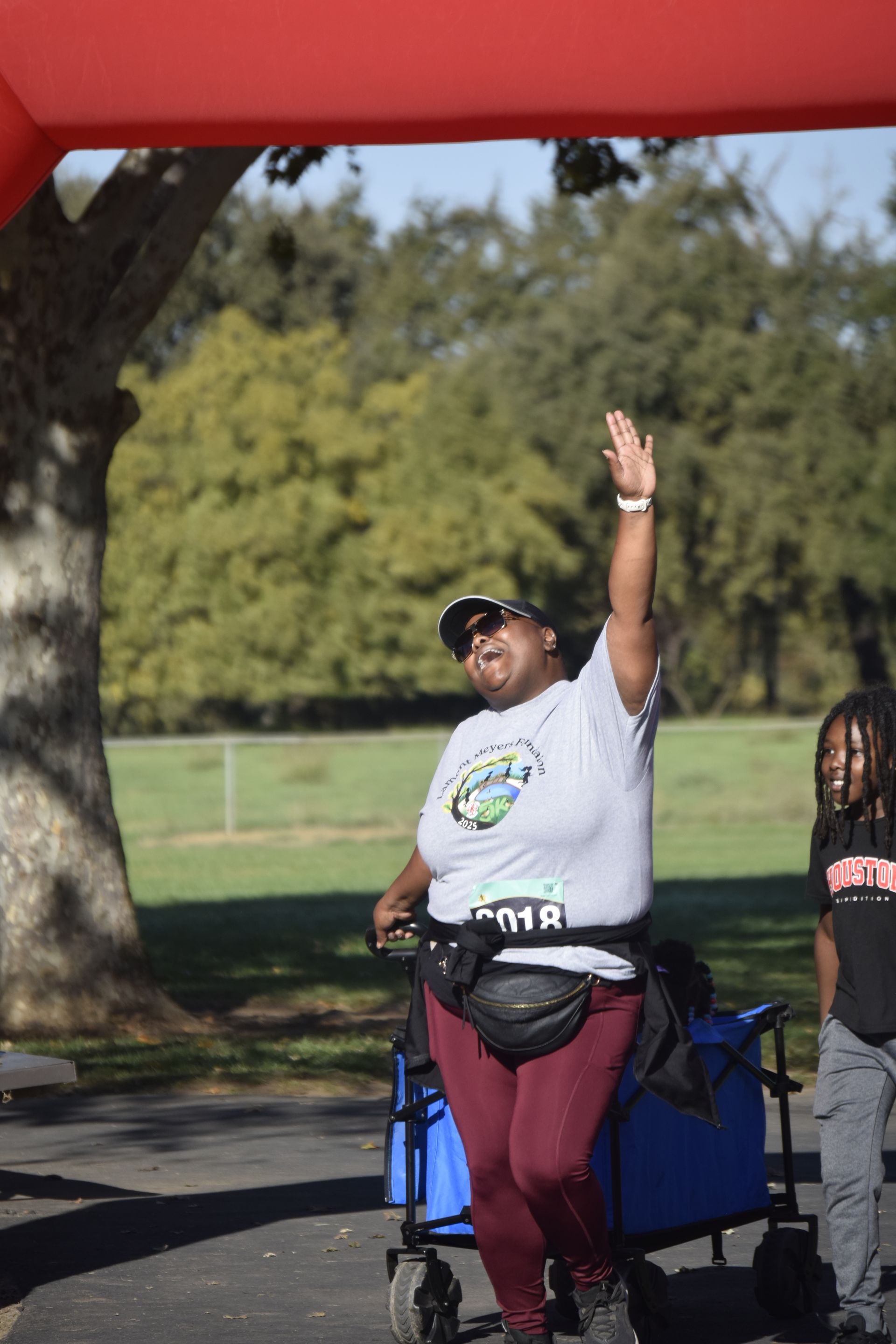 Woman waving with arm raised, crossing finish line of outdoor event, wearing race bib.