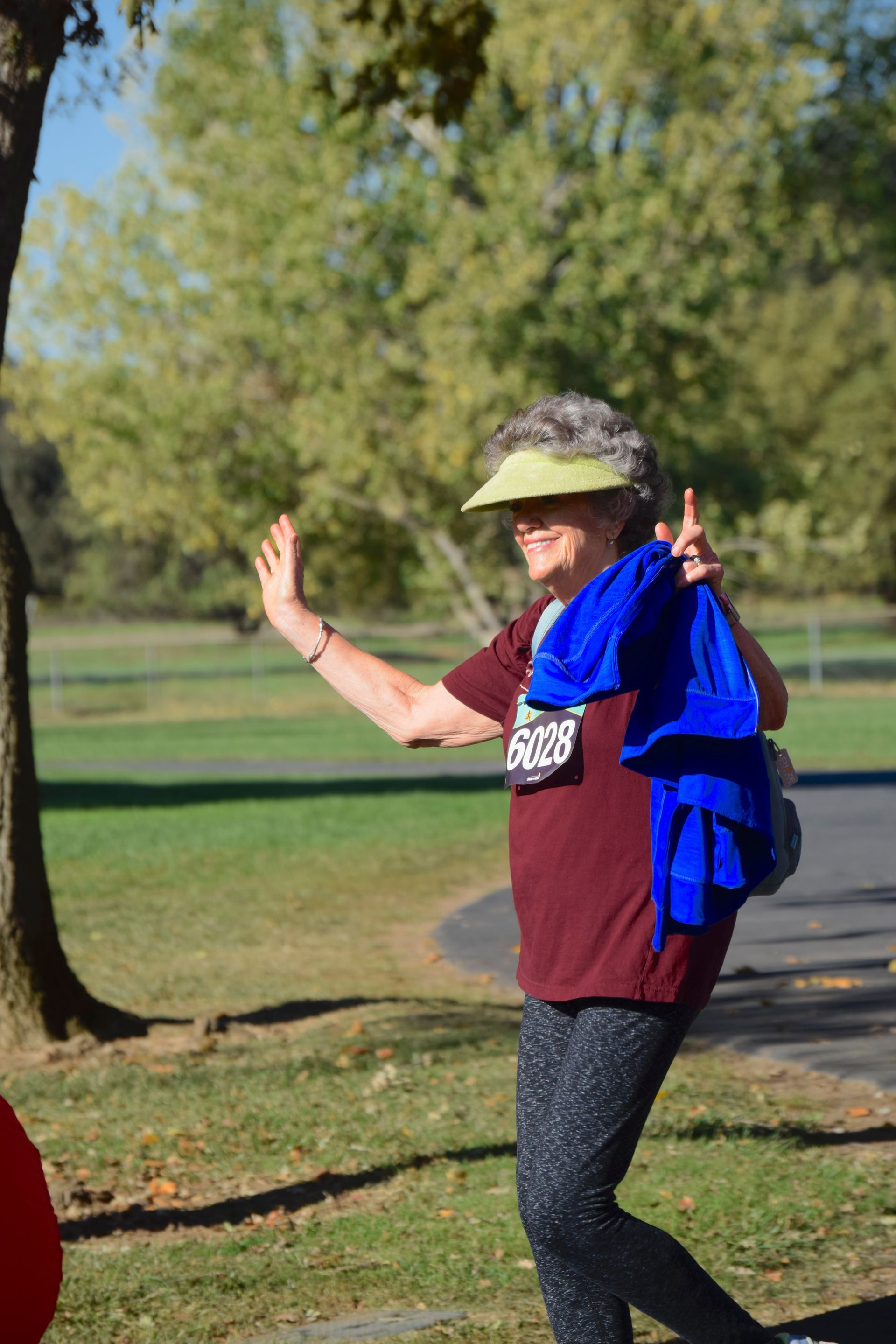 Woman waving, wearing visor and running bib, holding blue bag, standing in park.