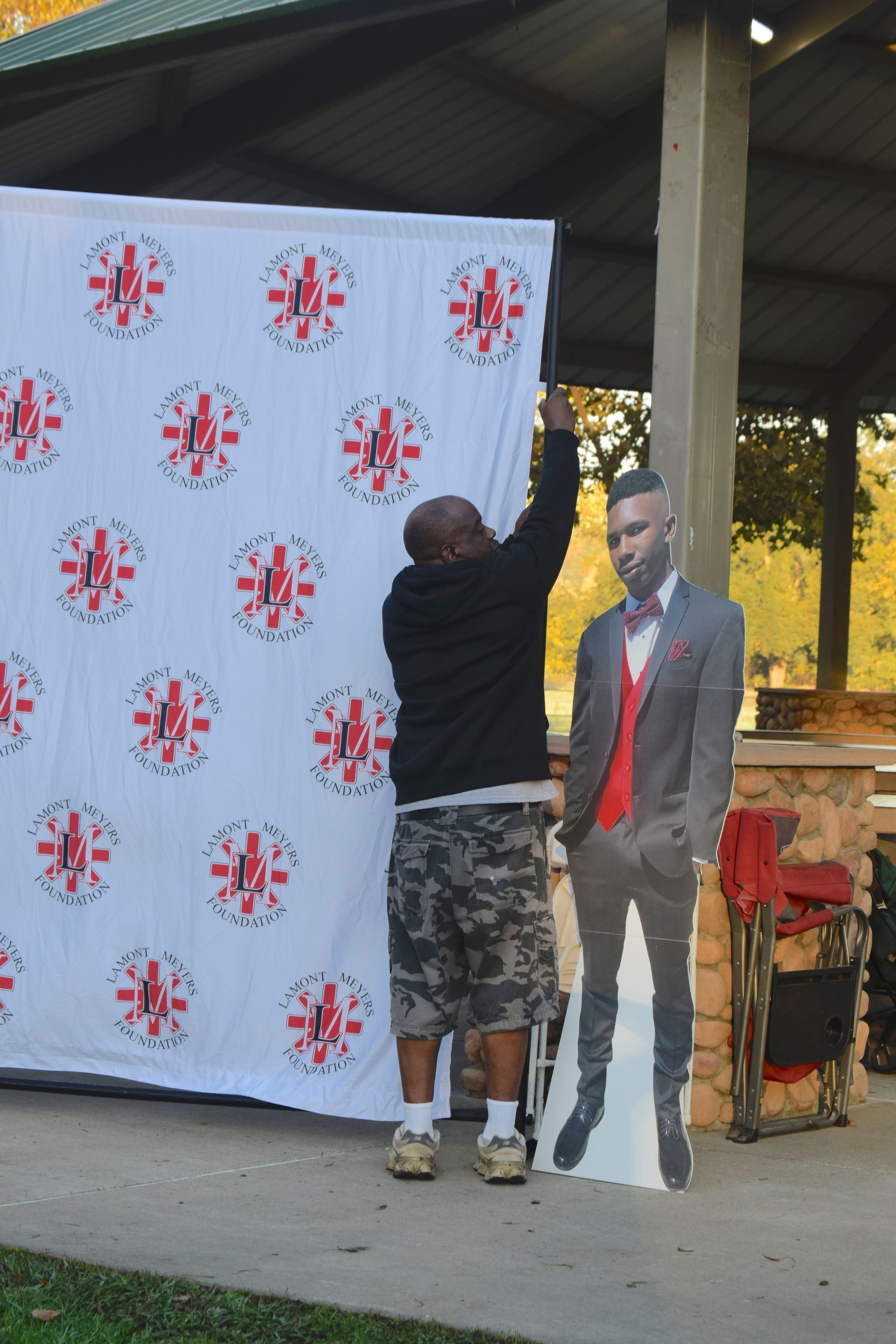 Man hanging a backdrop with a pattern of red and white designs next to a cardboard cutout of a man in a park.