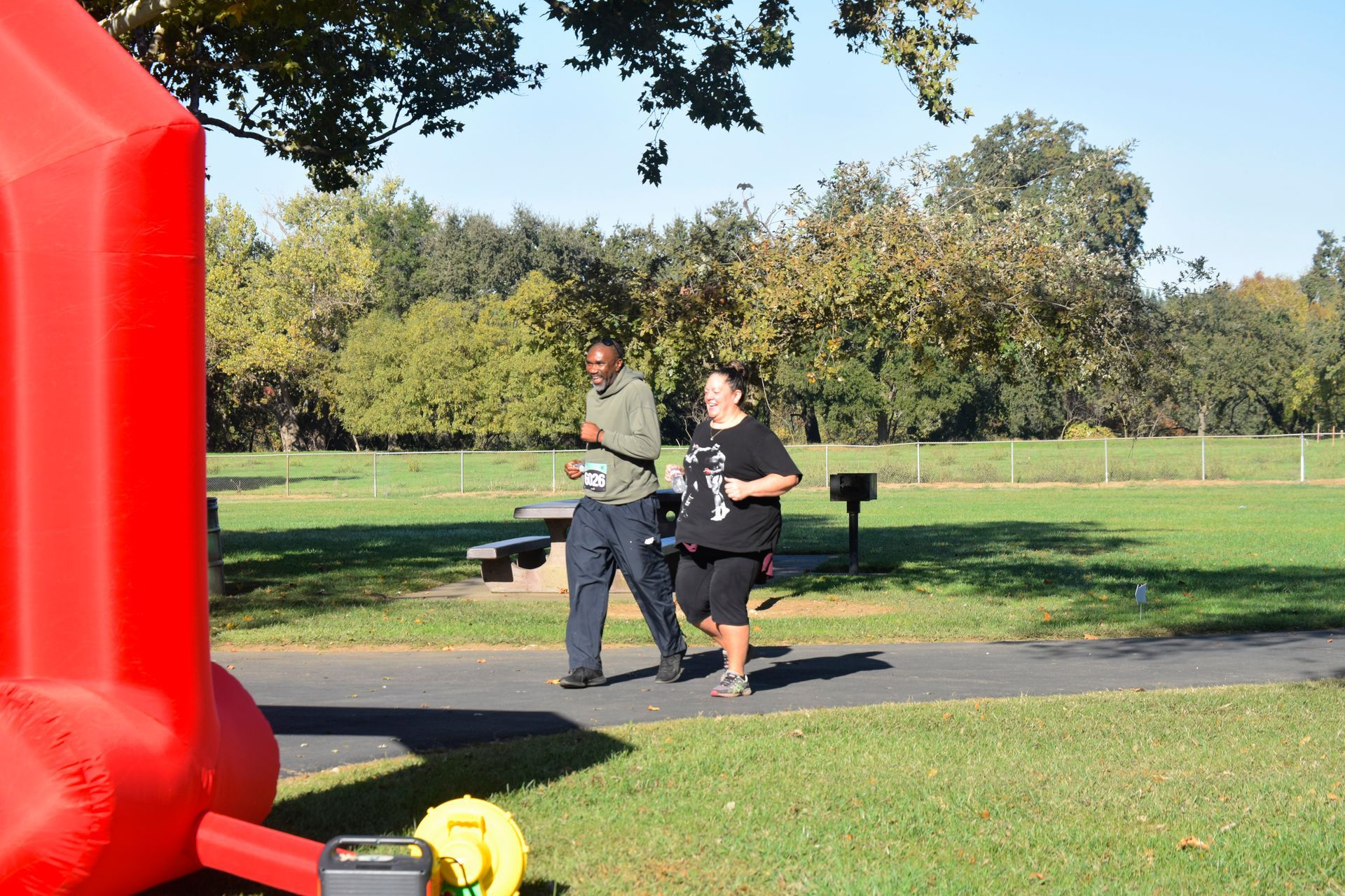 Two people running on a paved path in a park with green grass and trees; red inflatable on the left.