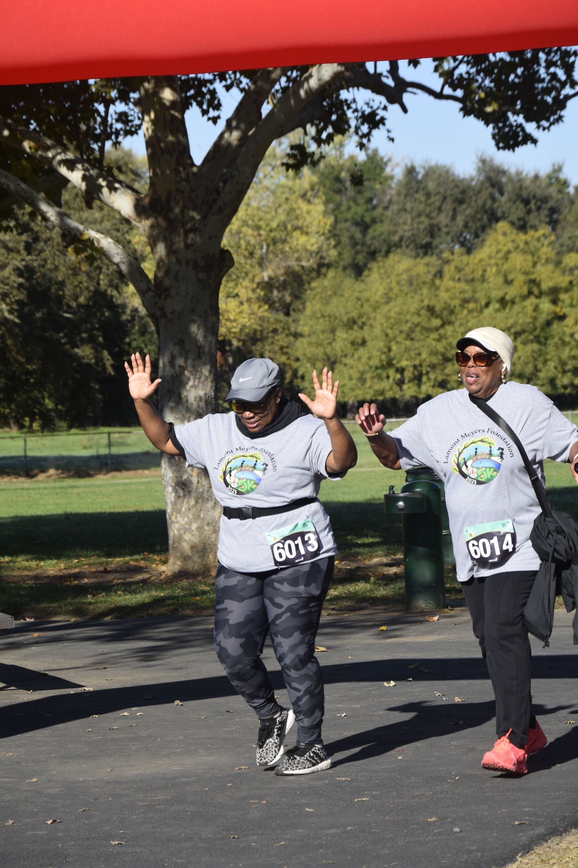 Two people crossing a race finish line. They are raising their hands and smiling, with a park setting in the background.