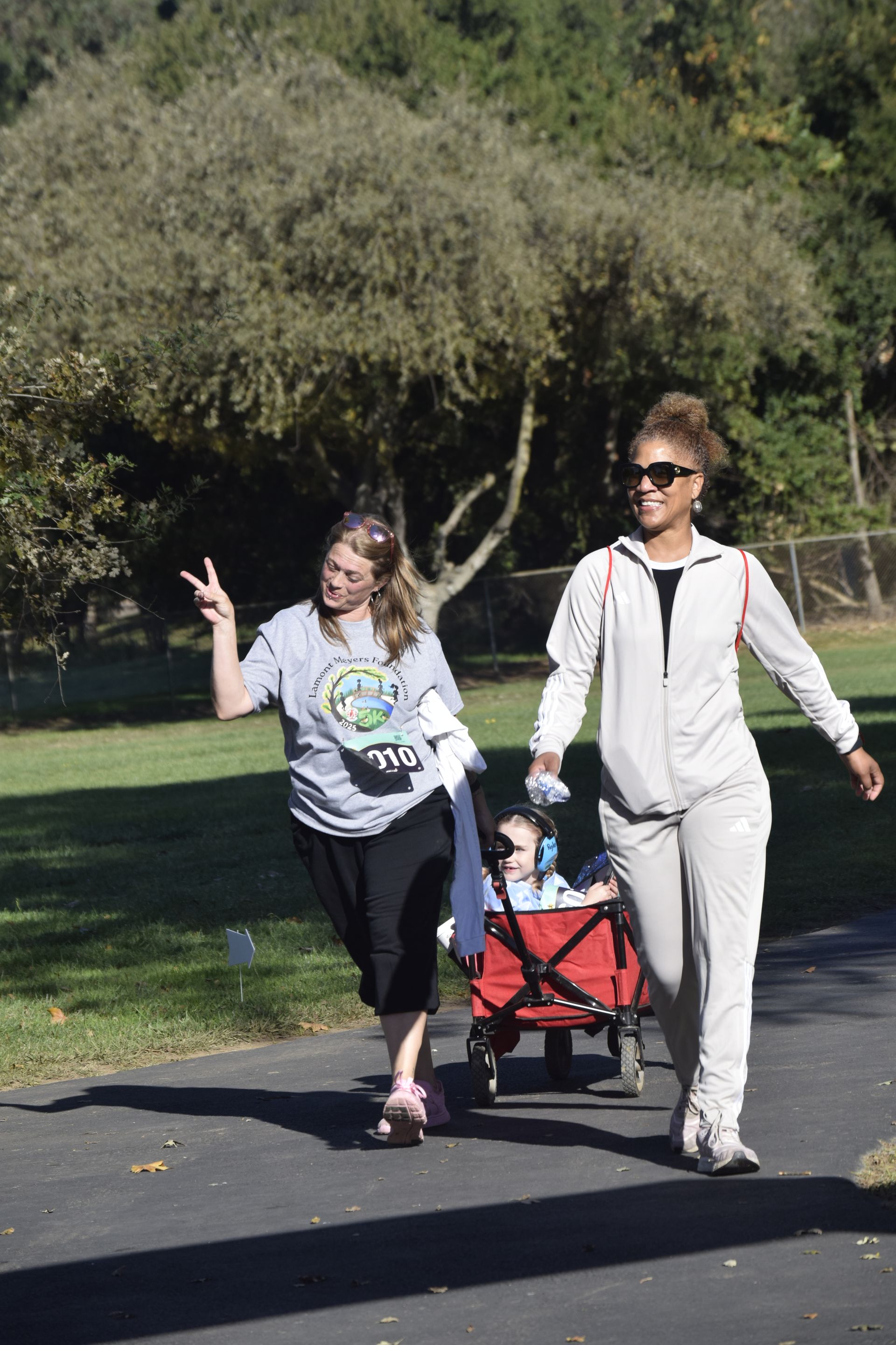 Two people walking on path, one pulling child in red wagon; trees in background, sunny day.