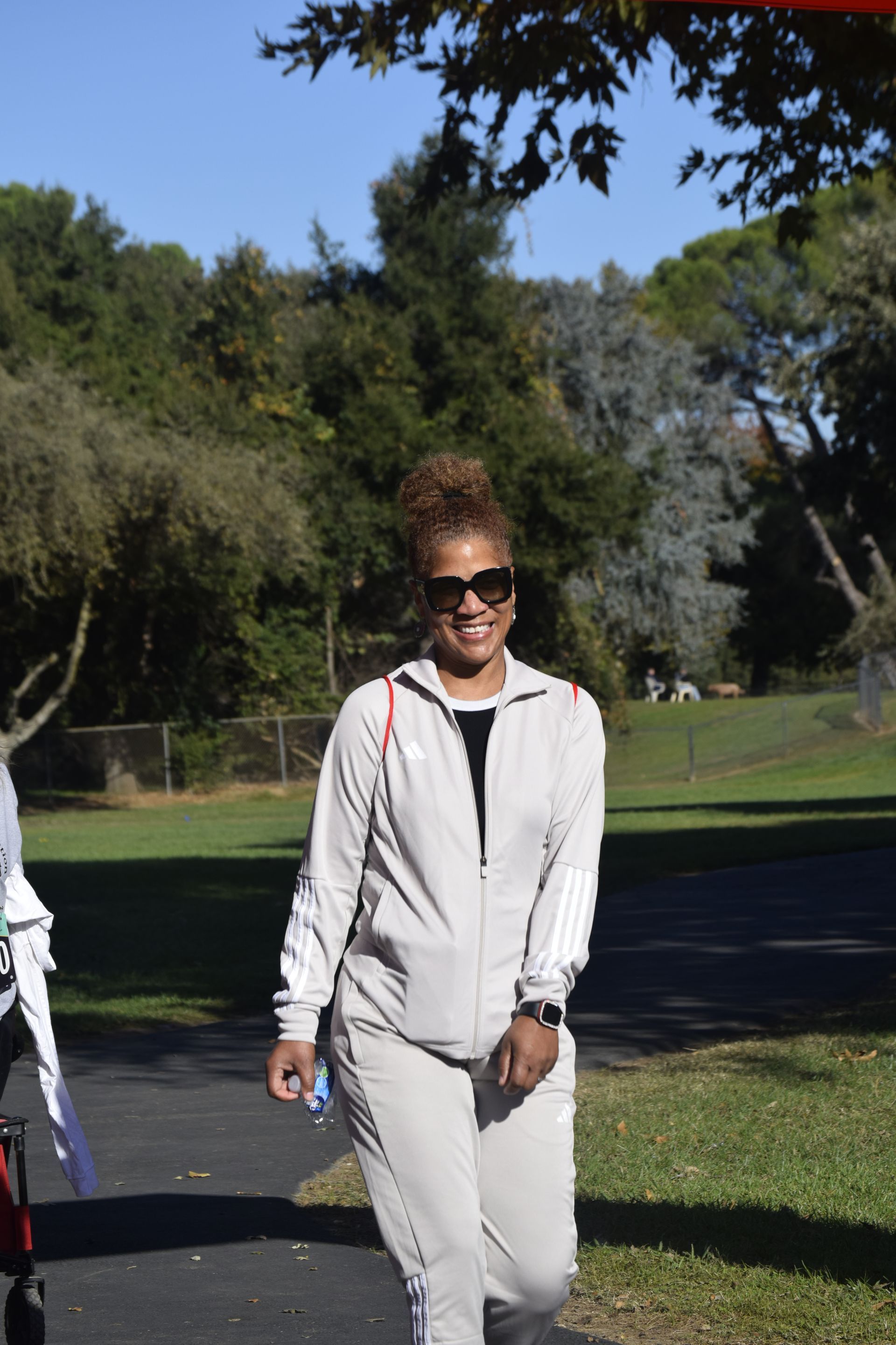 Woman in light-colored sweatsuit and sunglasses smiles walking outdoors. Trees and grass in background.