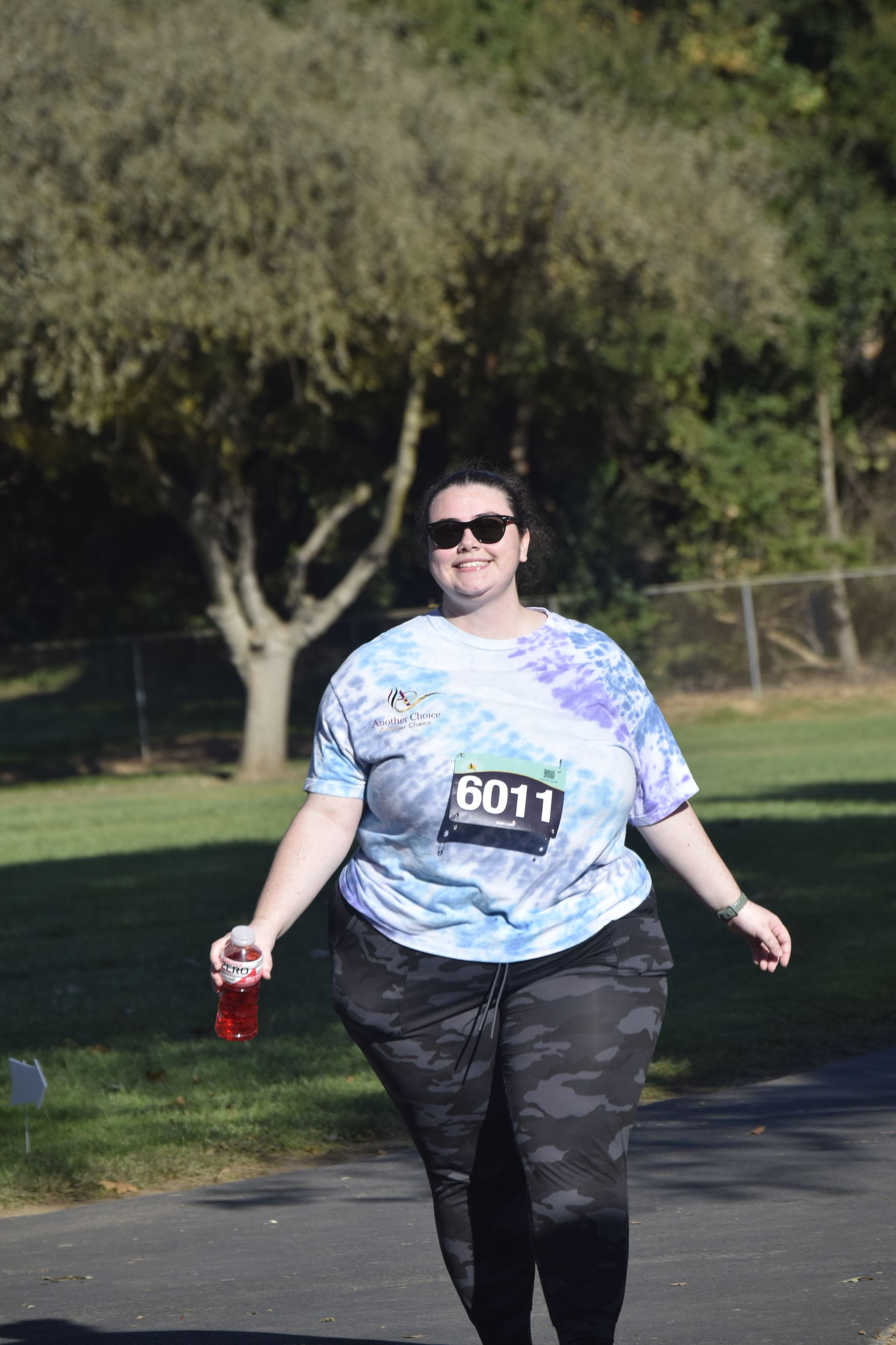 Woman in tie-dye shirt and camo leggings, running a race, smiling, holding a drink, sunny day.