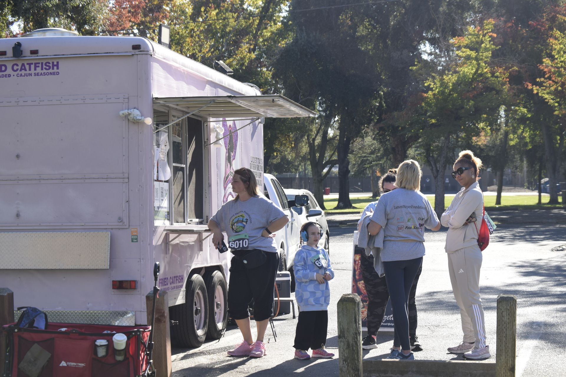 People line up at a pink food truck in a park on a sunny day.