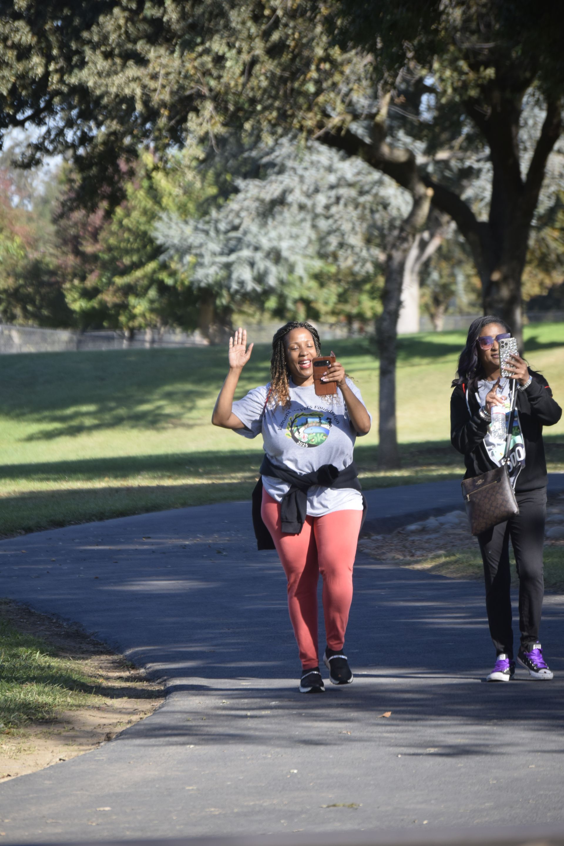 Two people on a paved path in a park. One waves, holding a phone, the other photographs. Sunny day.