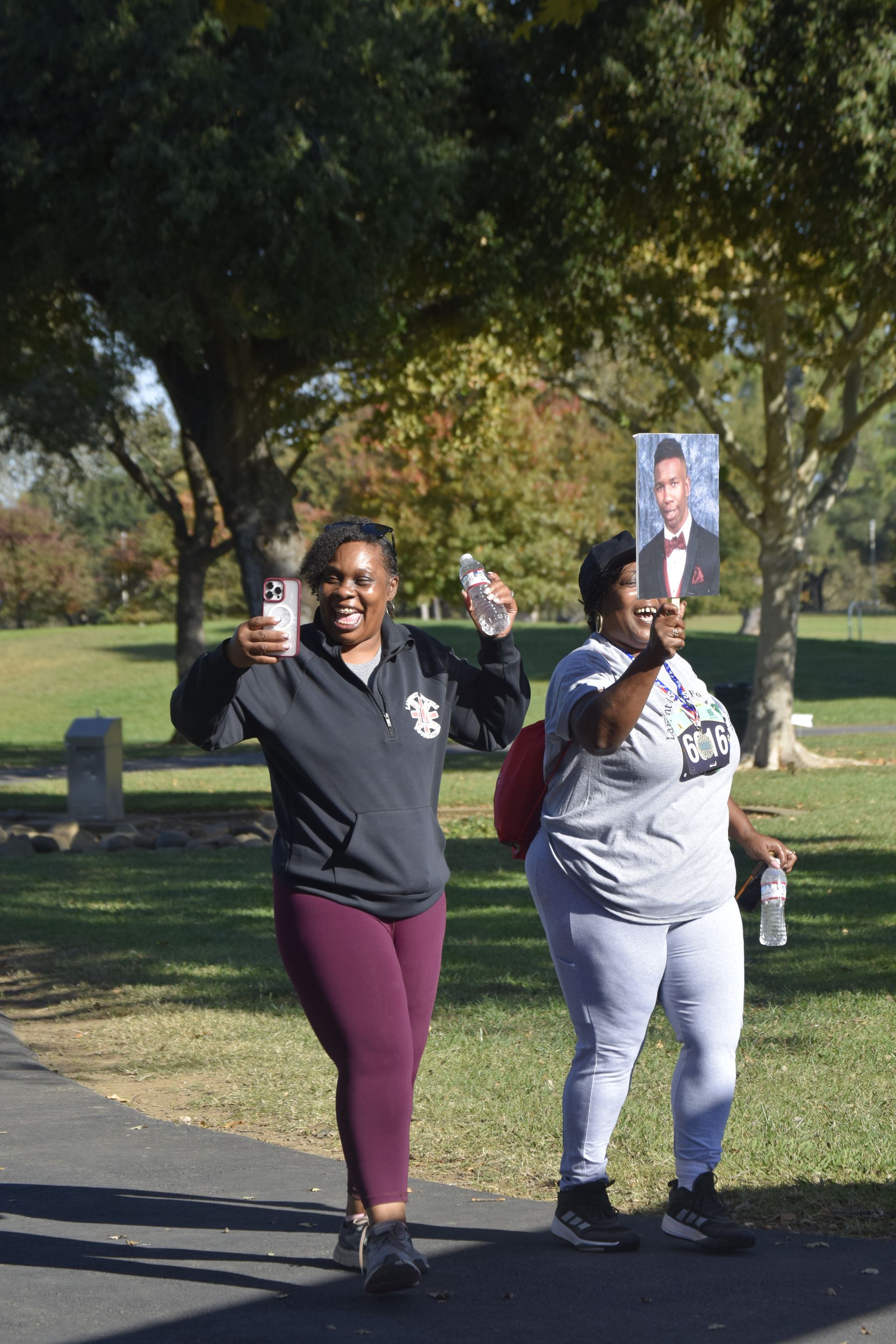 Two people walking in a park, one holding a photo, both smiling and holding water bottles.