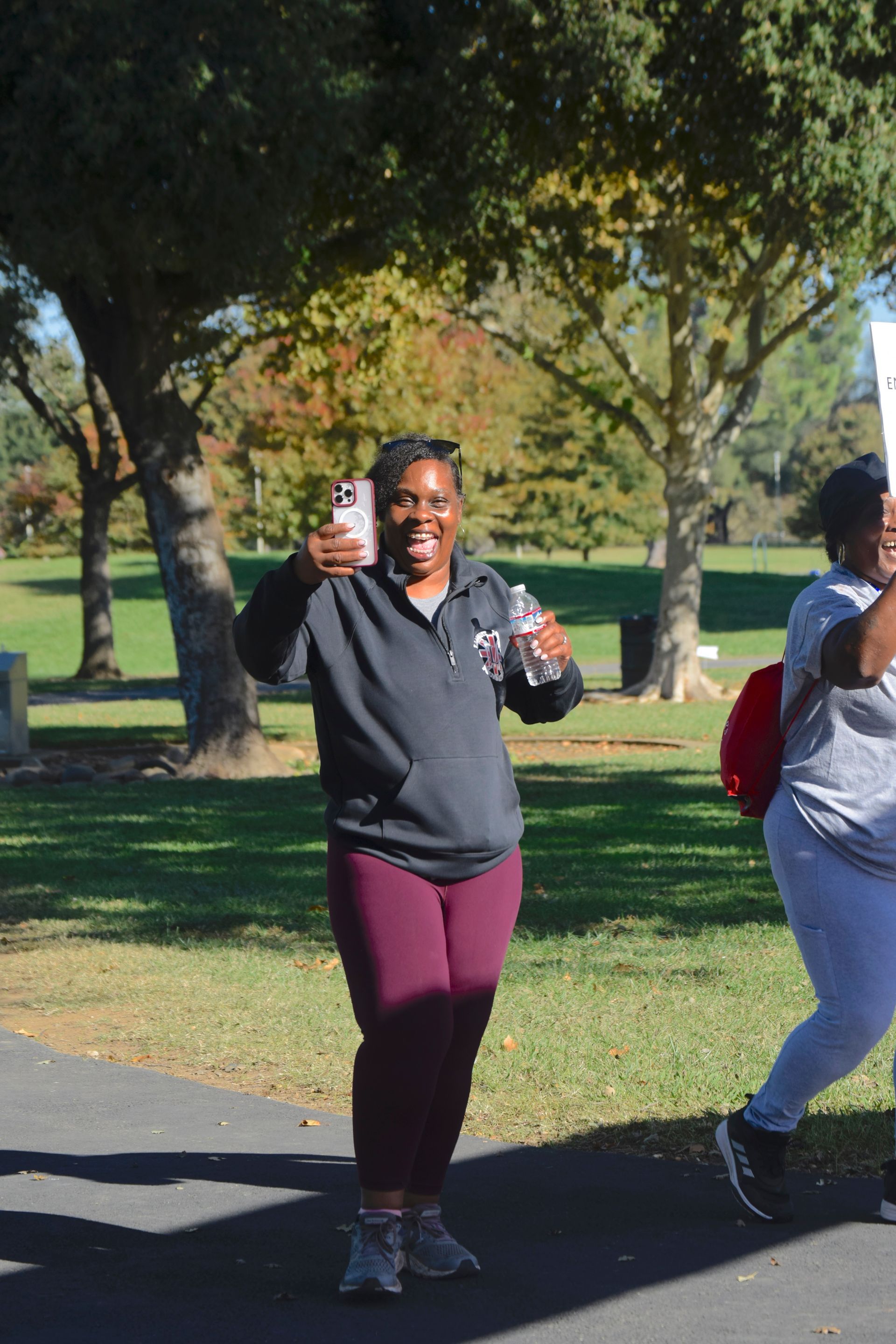 Woman in maroon leggings and a black hoodie holding a phone and smiling outdoors, walking on a path.