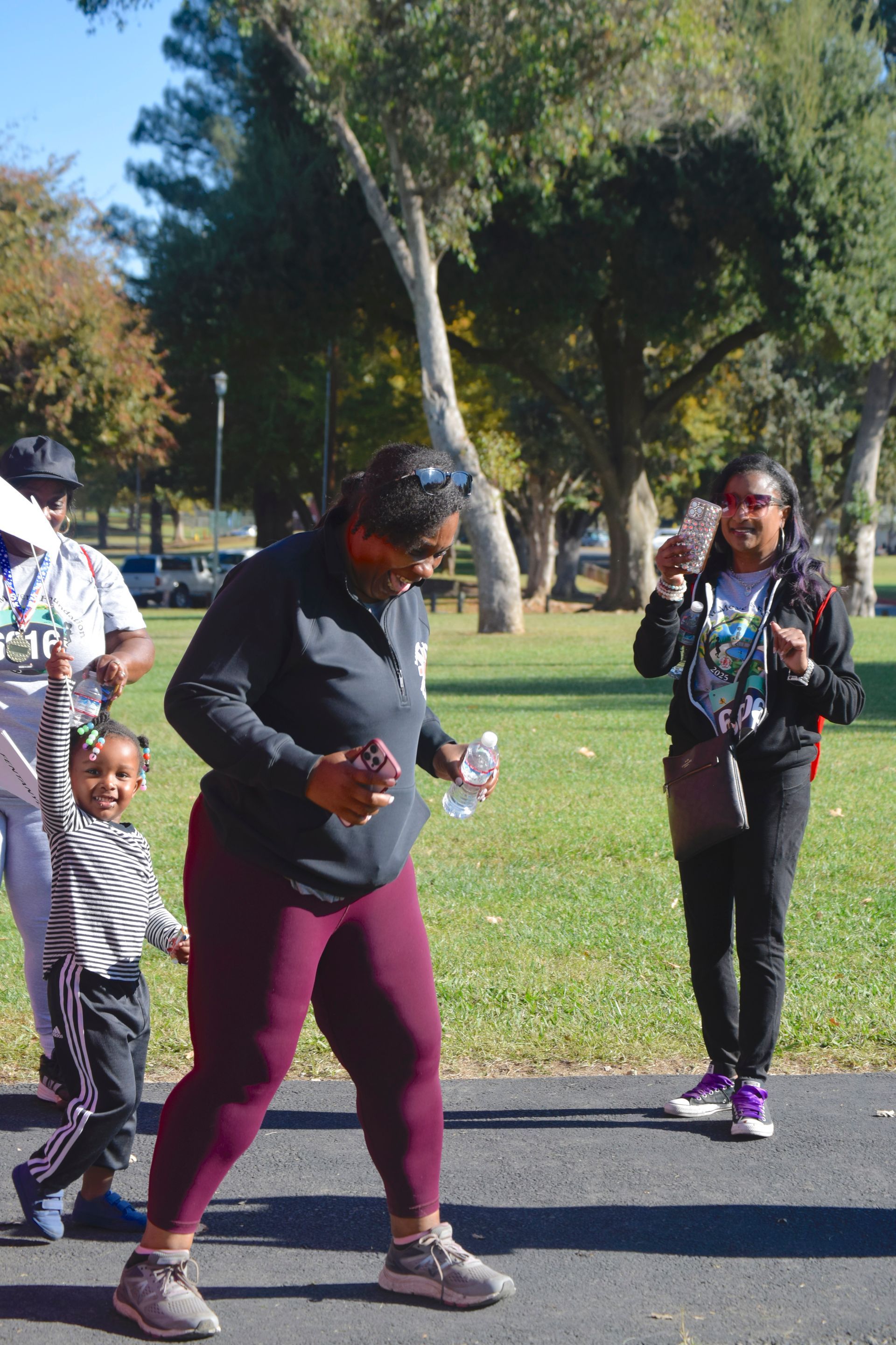 People walking outdoors in a park; one person dancing and holding a water bottle, child raising arm.