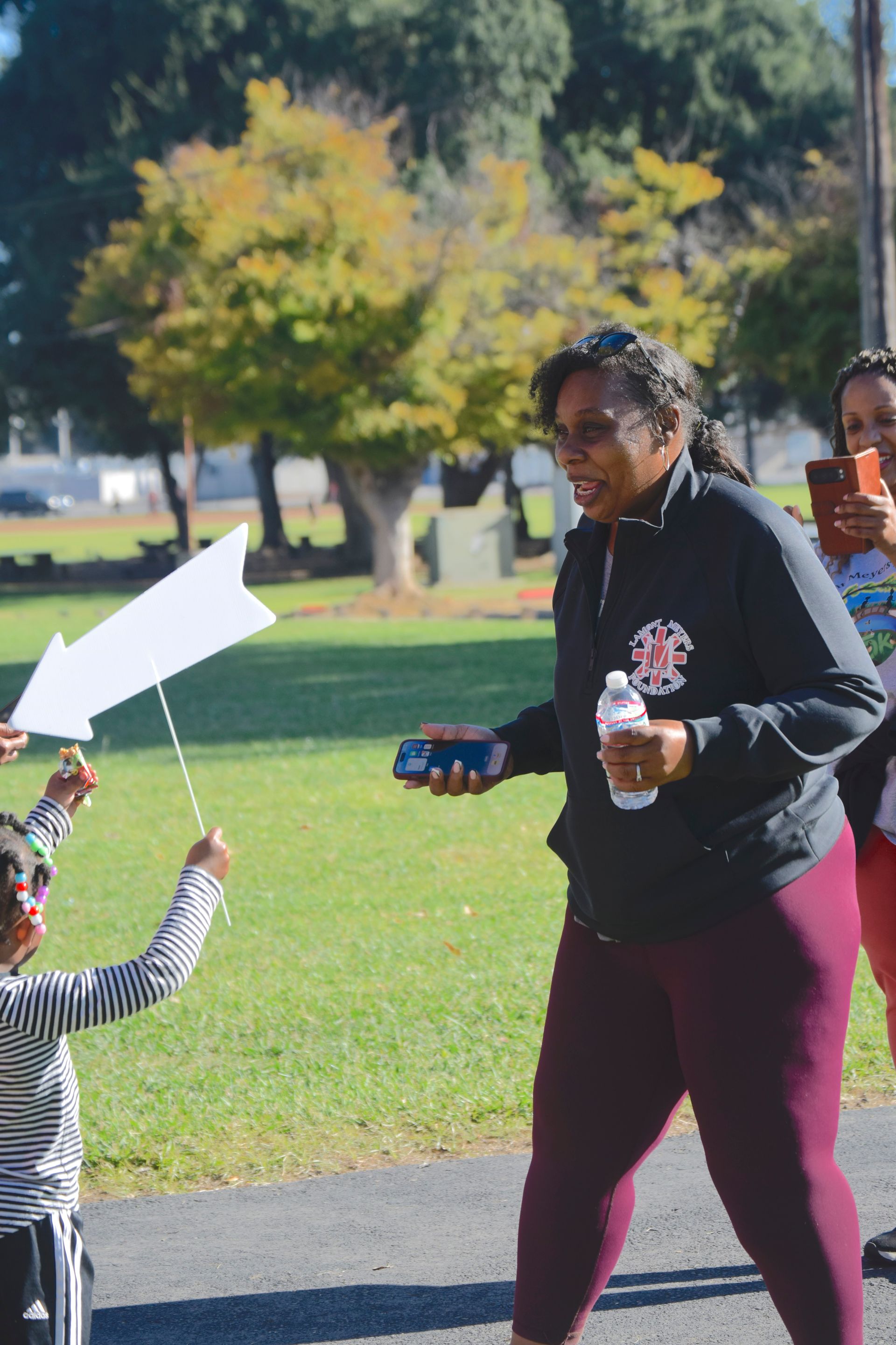 Woman holding water bottle and small container, smiling, with a child holding an arrow sign. Park setting.