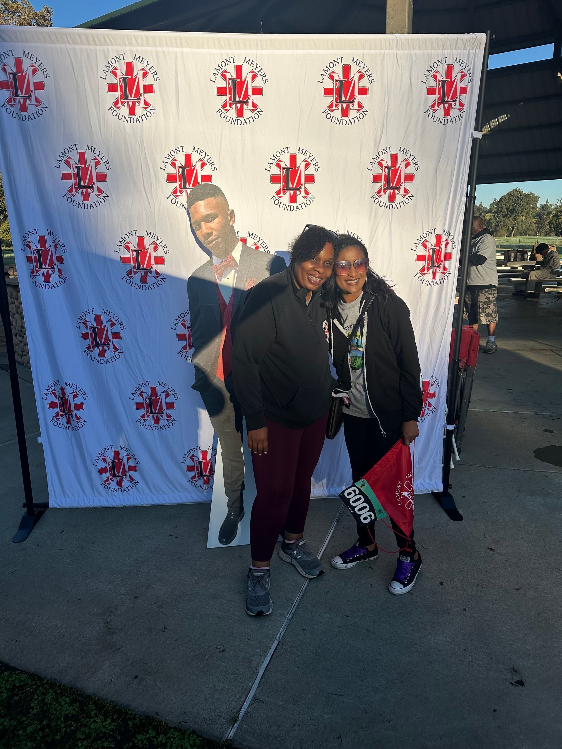 Two people smile posing beside a backdrop with a cutout figure. Outdoor setting, daylight.
