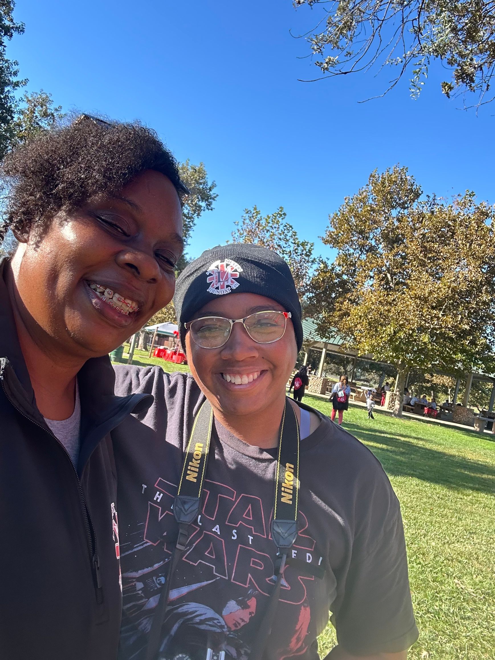 Two people smiling outdoors, near trees. Sunny day, one wearing hat and glasses.